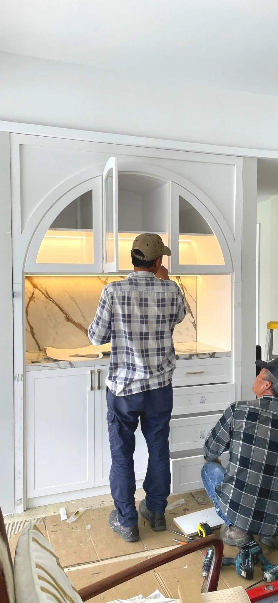 Two workers installing white cabinets with marble backsplash in a kitchen. One standing and the other kneeling, surrounded by tools and construction materials.