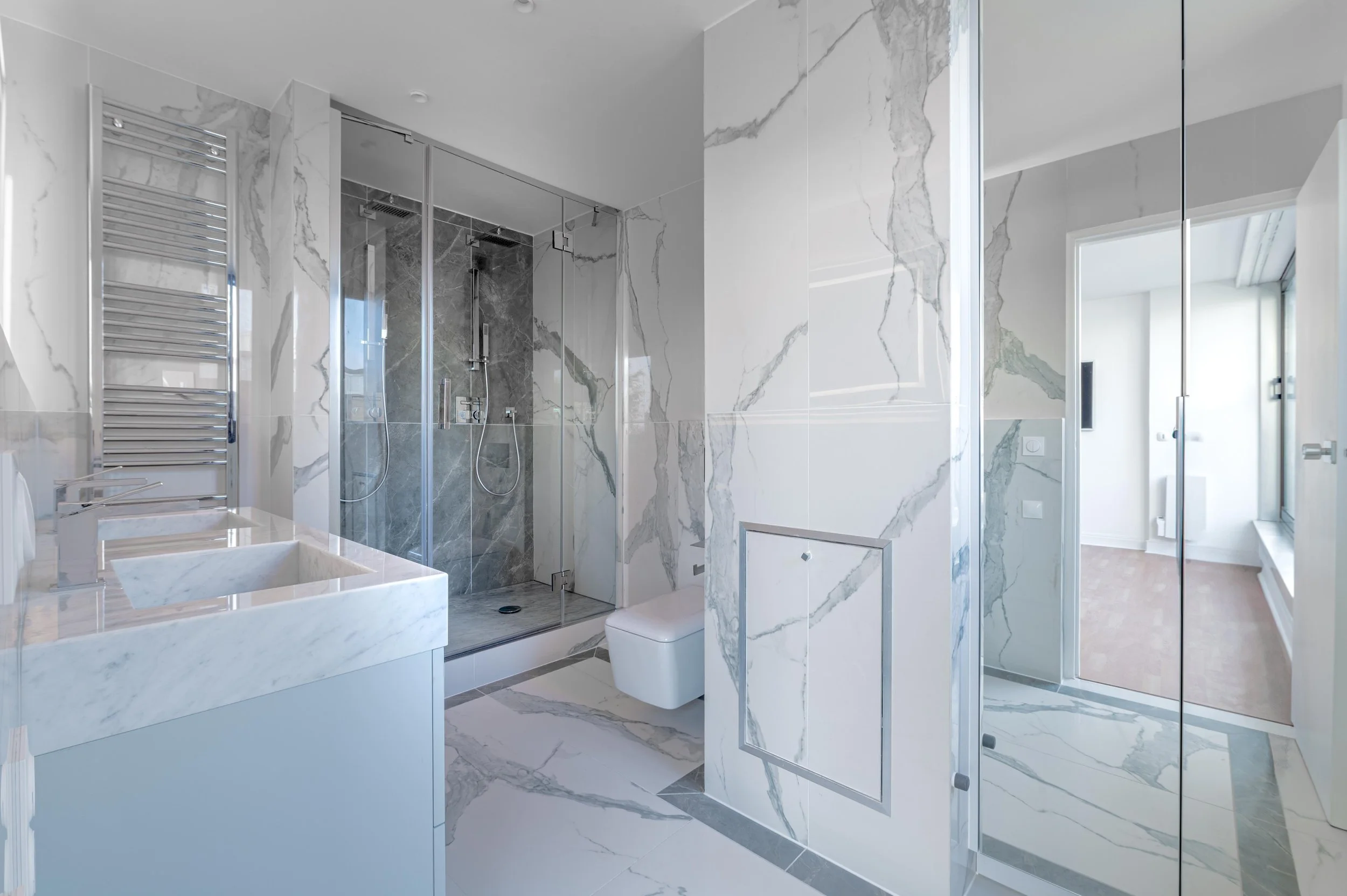 Salle de bain moderne avec murs et sol en marbre, douche en verre, lavabo en marbre, et toilette blanche. Un espace lumineux avec une grande fenêtre.