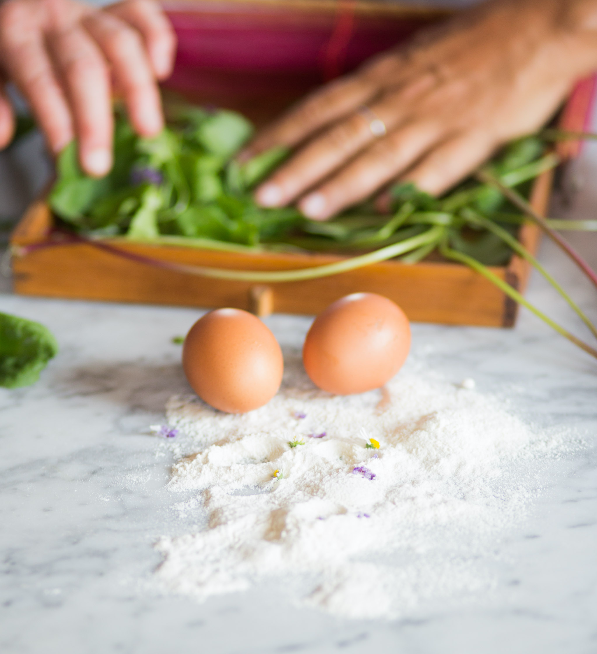 Close-up of three brown eggs on a white powdery surface with small flowers, with blurred green leaves and a wooden box with green and purple vegetables in the background.