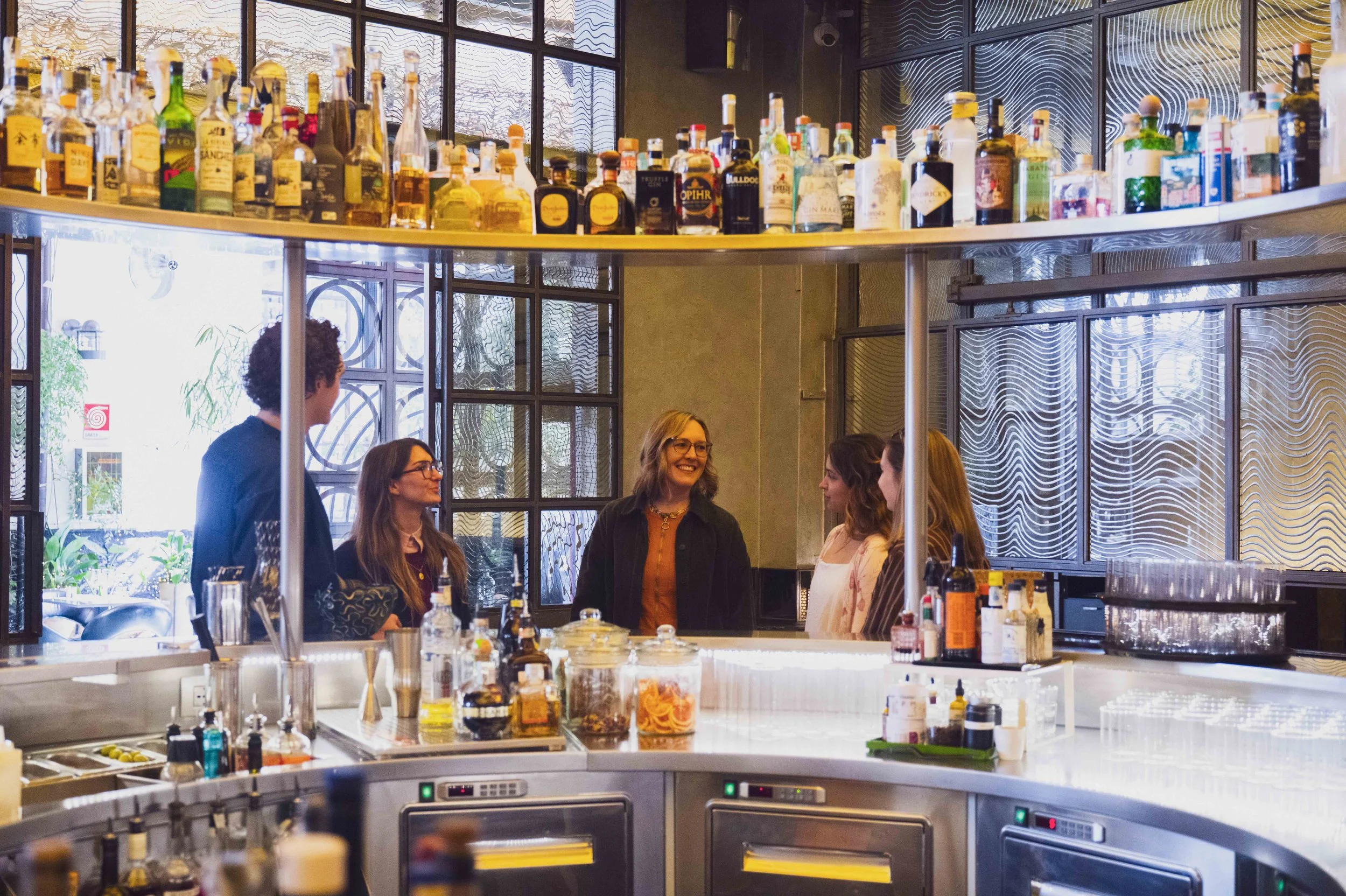 A group of five women talking in a bar or restaurant with shelves of liquor bottles behind them.