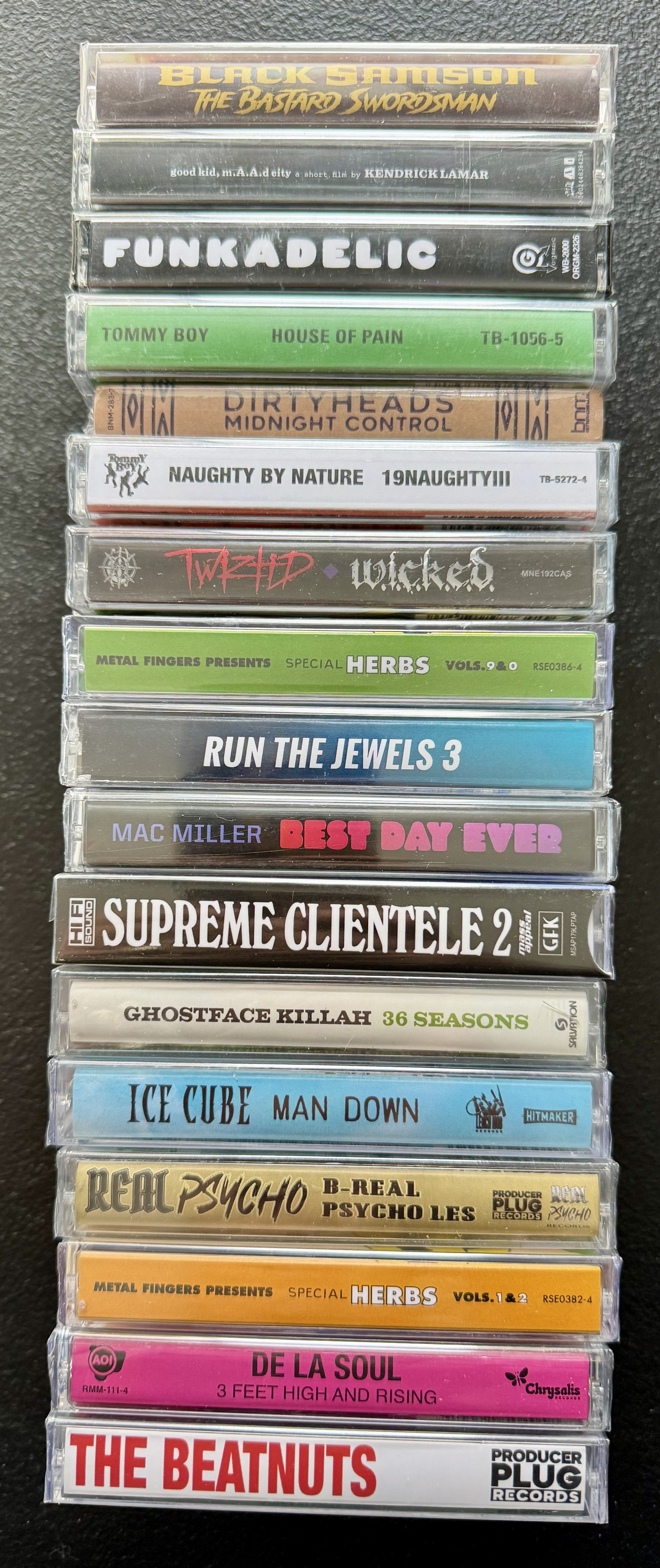 Stack of cassette tapes featuring various musical artists and albums, including Black Samson, Kendrick Lamar, Funkadelic, Tommy Boy, Dirty Heads, Twiztid, Herbs, Run the Jewels, Mac Miller, The Beatnuts, Ice Cube, Psycho, De La Soul, and The Beatnuts.