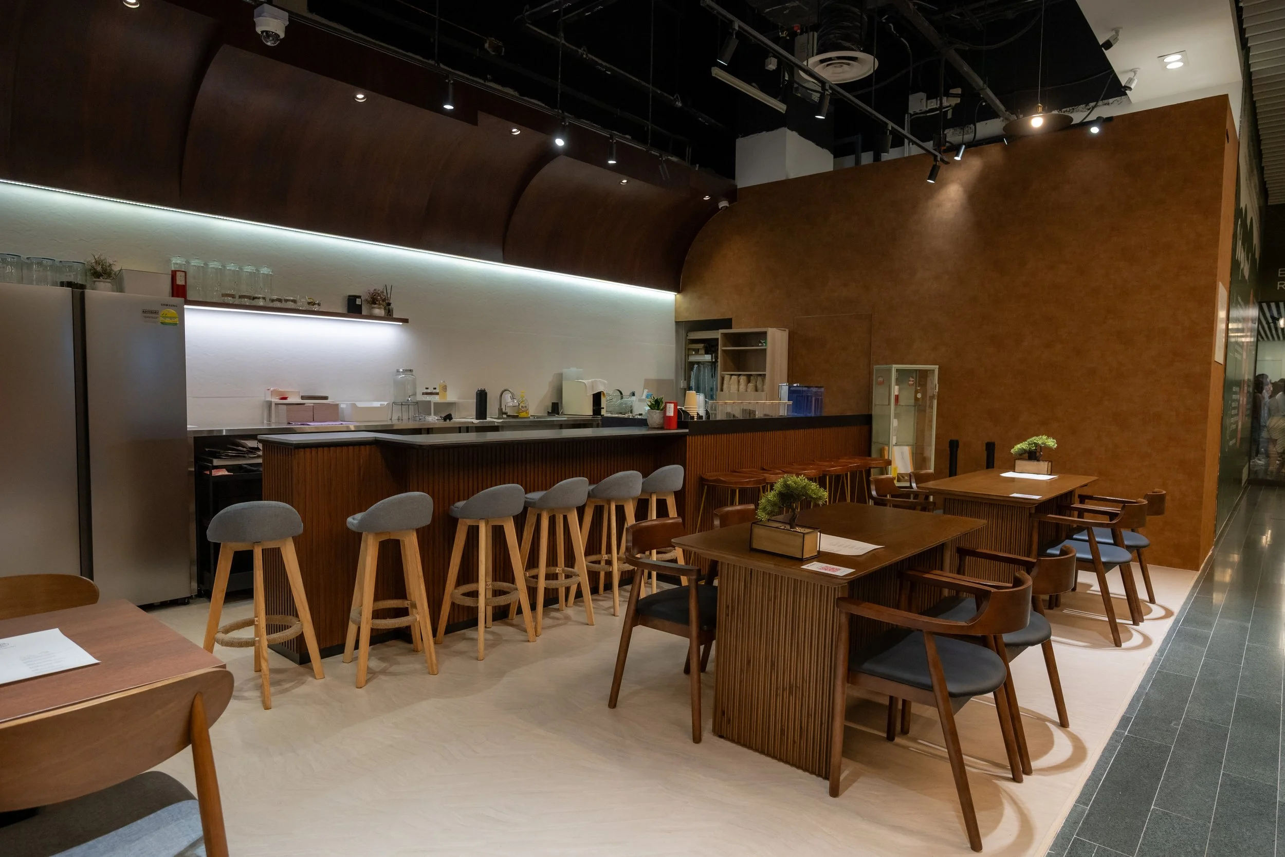 Interior of a modern cafe or restaurant with wooden tables and chairs, a bar counter with bar stools, and a kitchen area in the background.