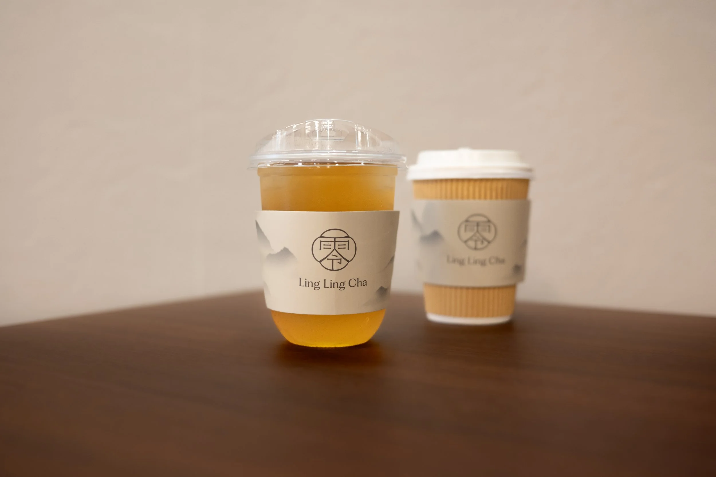 Two beverages, one in a clear plastic cup with a dome lid and the other in a paper cup with a lid, placed on a wooden surface with a plain background. Both cups have labels reading 'Ling Ling Cha' and a stylized logo.