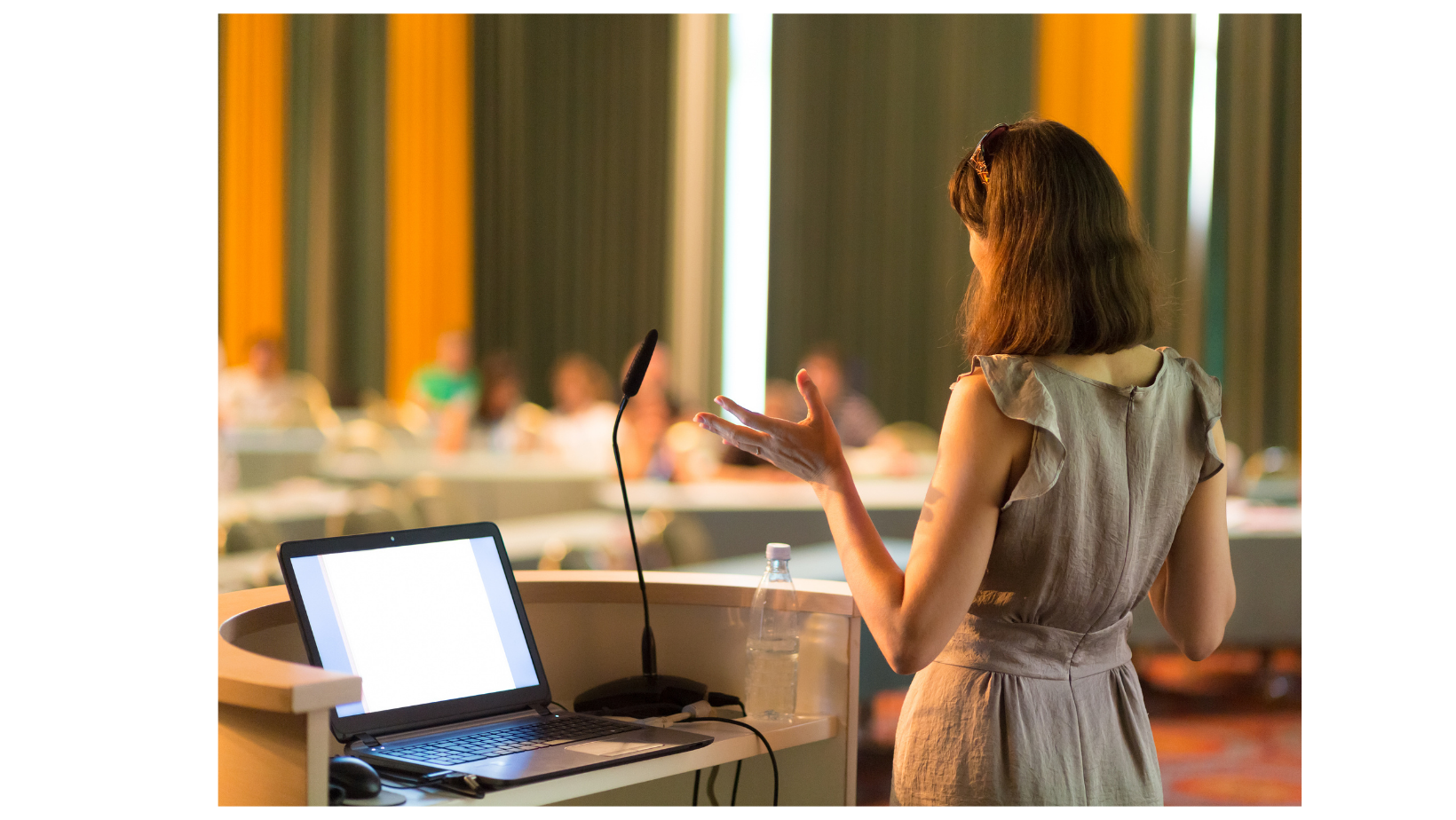 プレゼンテーション中の女性の後ろ姿と、開いたノートパソコン。テーブルにはマイクと水のボトルがある。