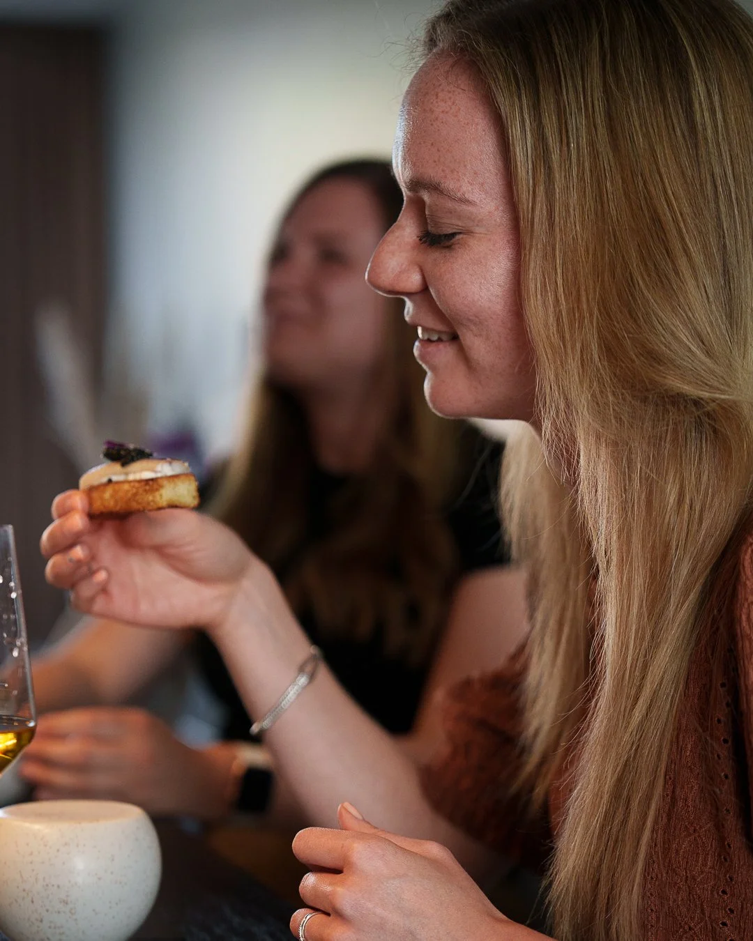 Person holding a small toast with topping, smiling, sitting with another person, and a wine glass.