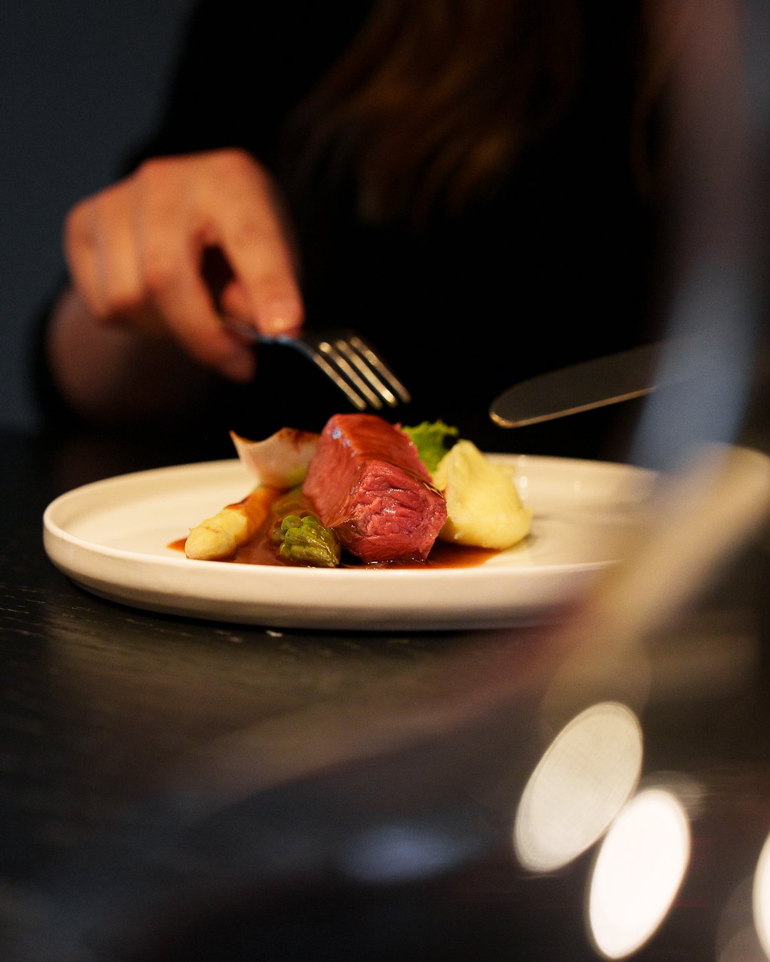 Person cutting into a cooked steak on a plate with asparagus, garnish, and sauce.