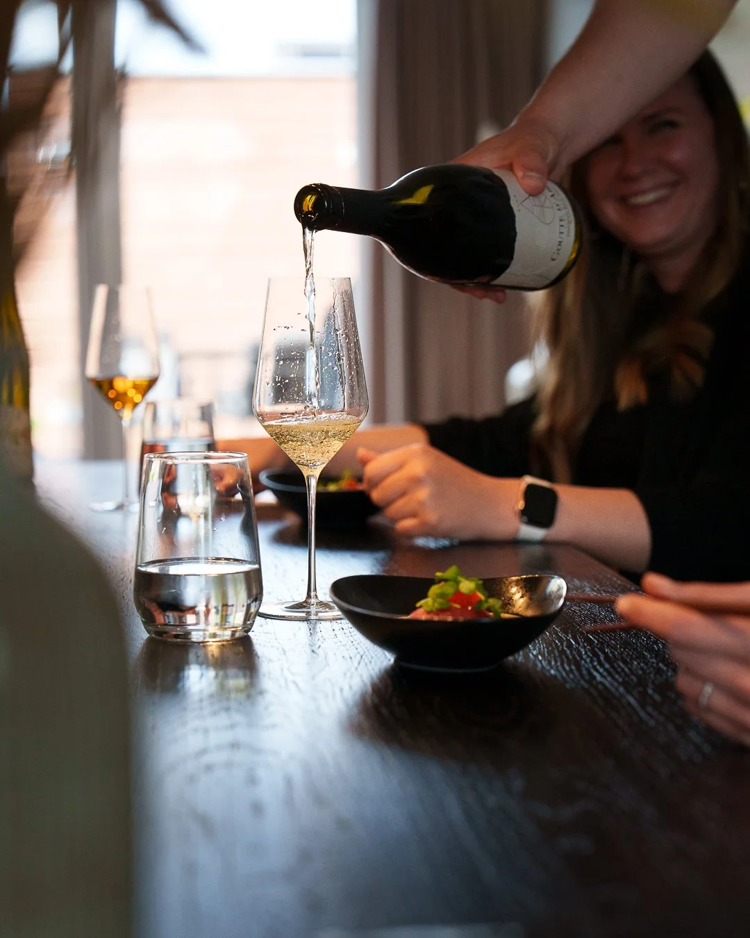 Person pouring wine into a glass at a dining table with food and other drinks, woman smiling in background.