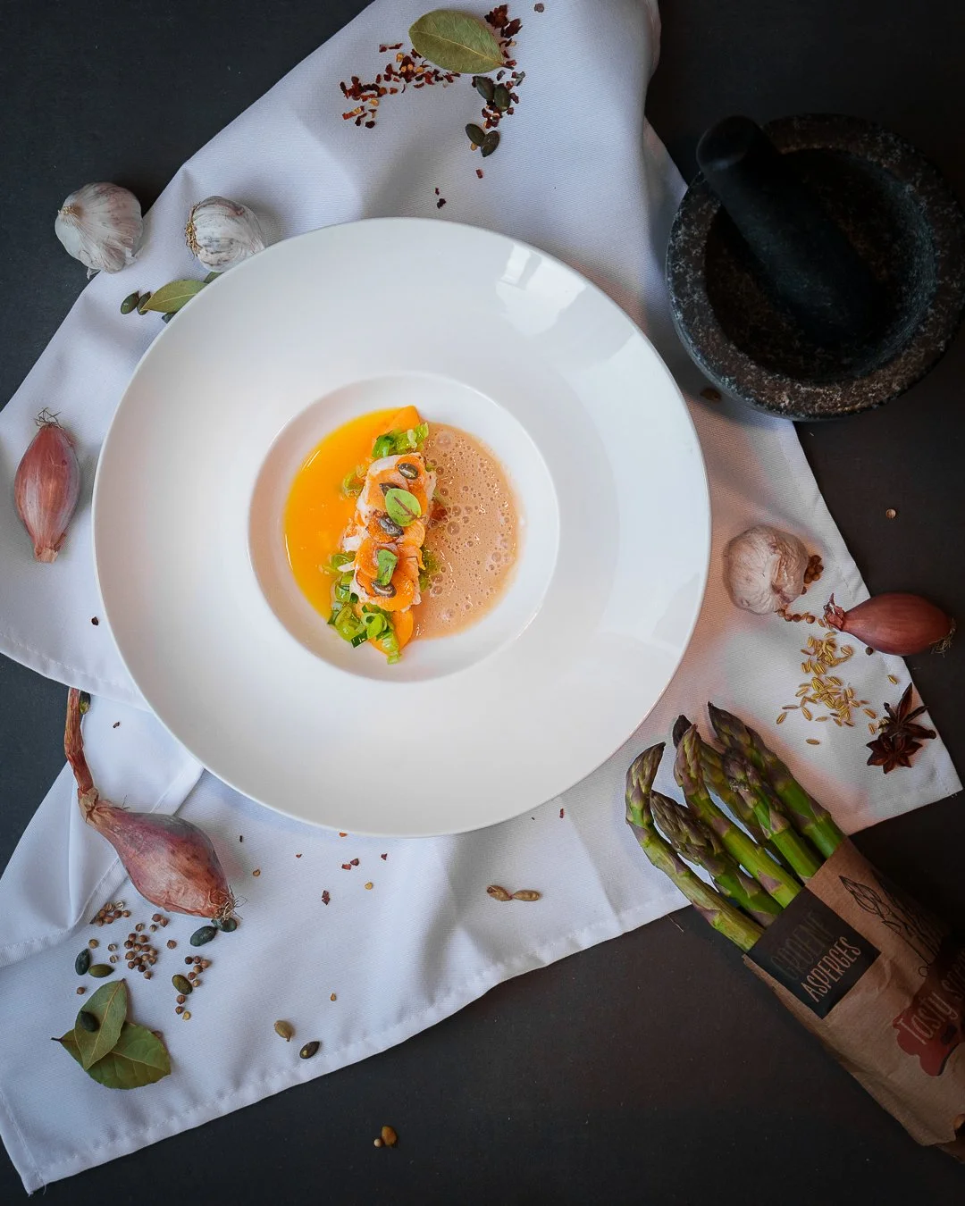 Plated gourmet dish with garnish, surrounded by garlic, shallots, spices, asparagus, and a mortar and pestle on a white cloth.
