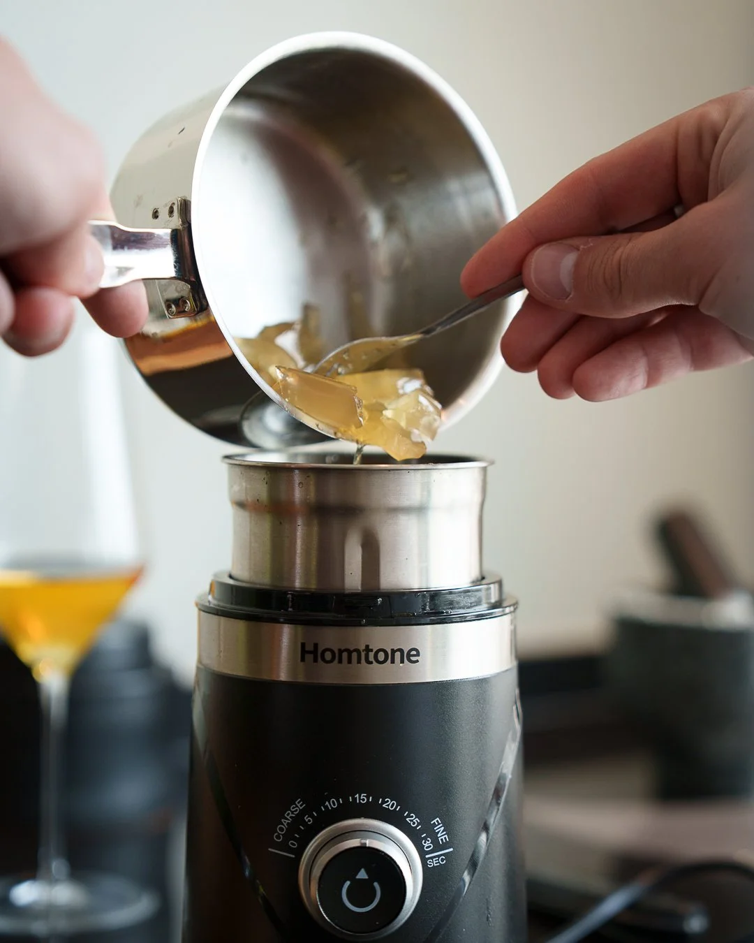 Close-up of hands using a spoon to transfer a gelatin-like substance from a metal pot into a Homtone appliance. A glass of amber liquid sits in the background.