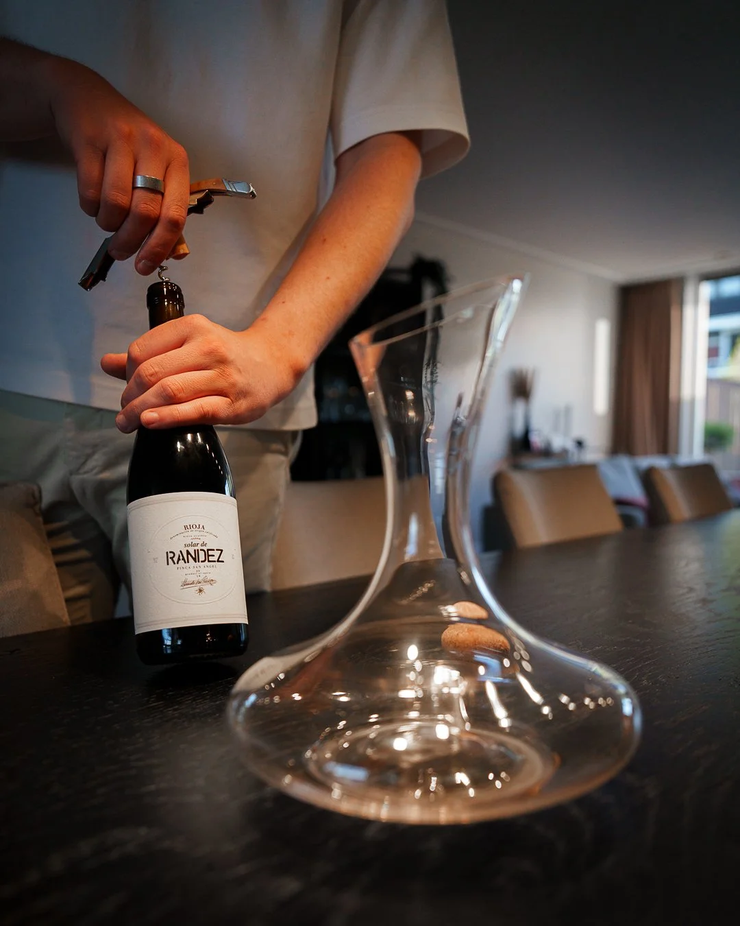 Person opening a wine bottle with a corkscrew next to a clear glass decanter on a dark table.