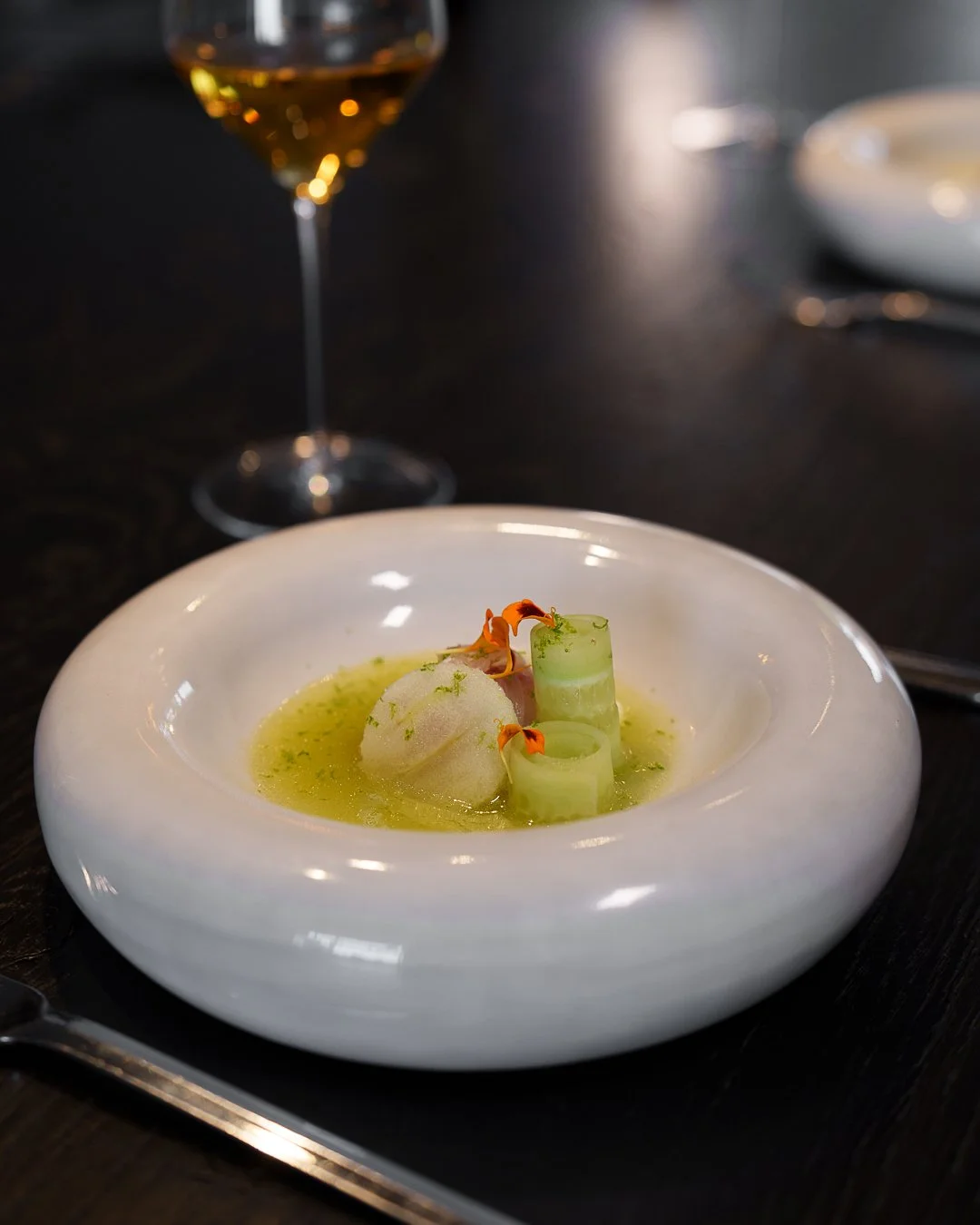 Gourmet dish in a white bowl with cucumber rolls, a quenelle of sorbet, and edible flowers, served with a glass of white wine on a dark table.