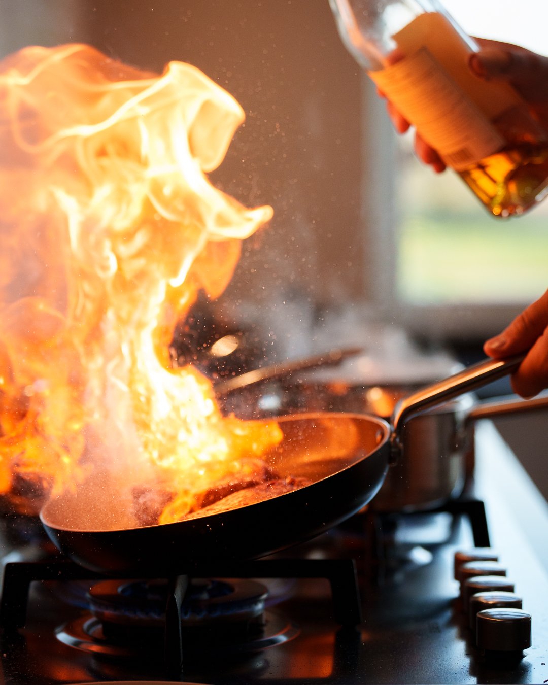 Person flambéing a pan of food on a stove with flames rising.
