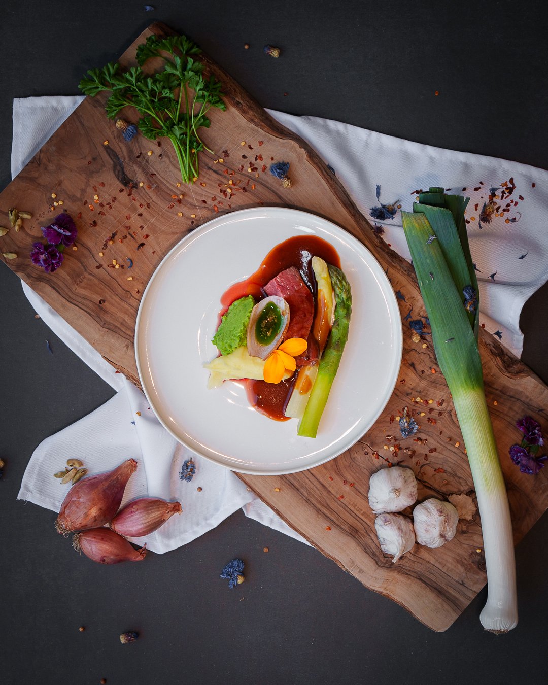 A gourmet dish plated on a white plate with beef, sauces, asparagus, and garnish, set on a wooden cutting board with a leek, garlic, and shallots nearby, adorned with herbs and flowers.