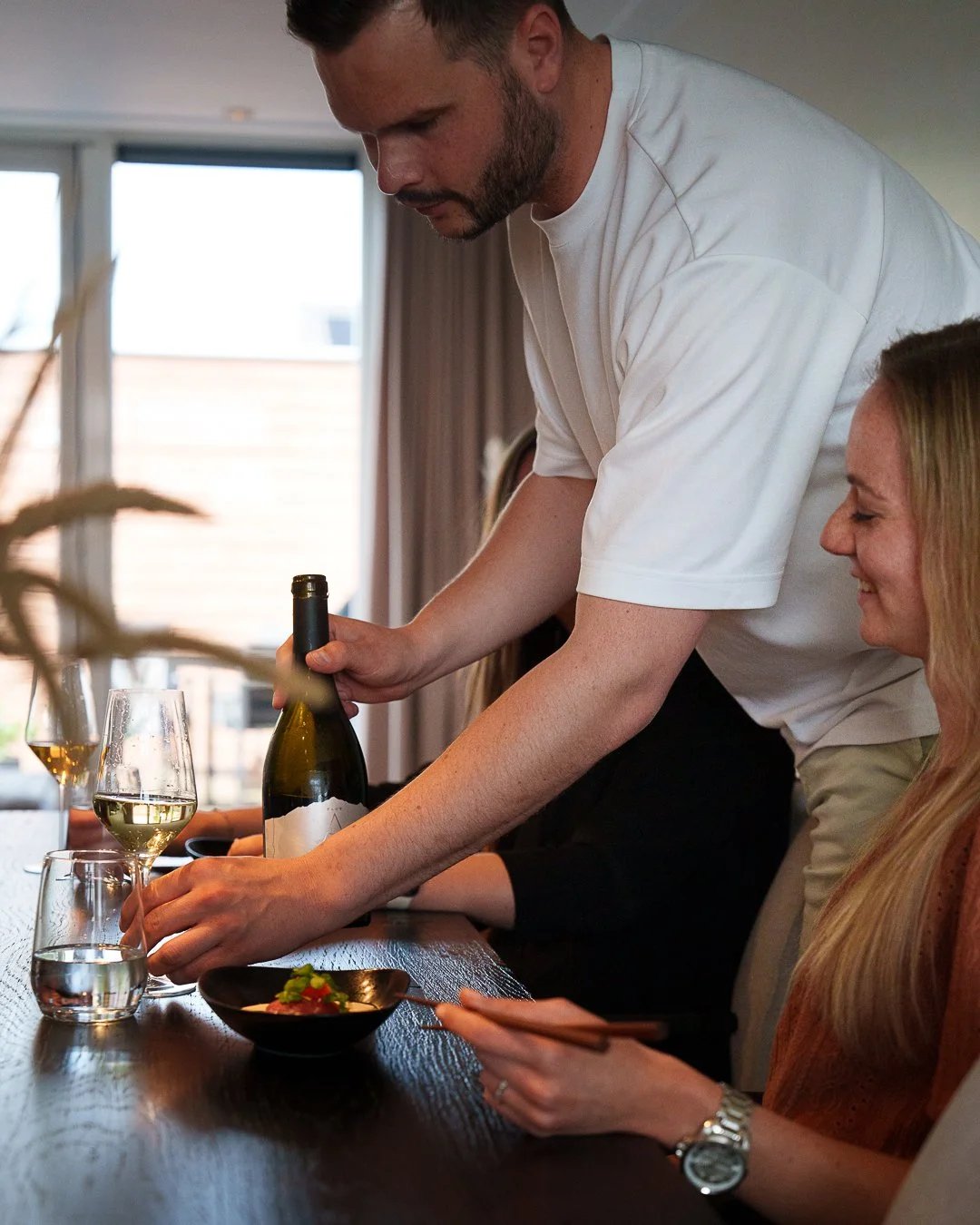Man pouring wine at a table with a woman sitting nearby, dining setup with glasses and a small bowl on a wooden table.