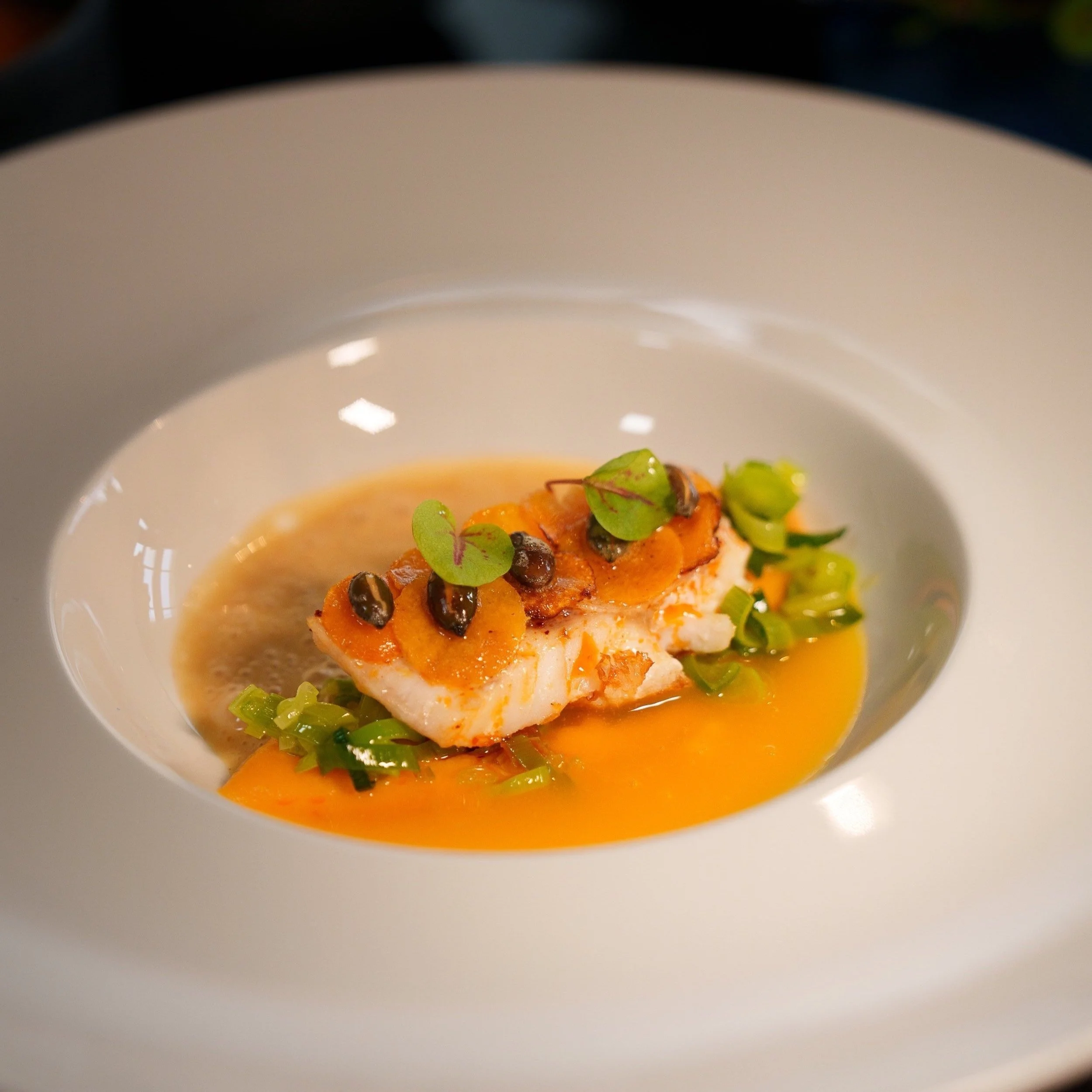Gourmet dish with fish filet, capers, microgreens, and orange sauce in a white bowl.