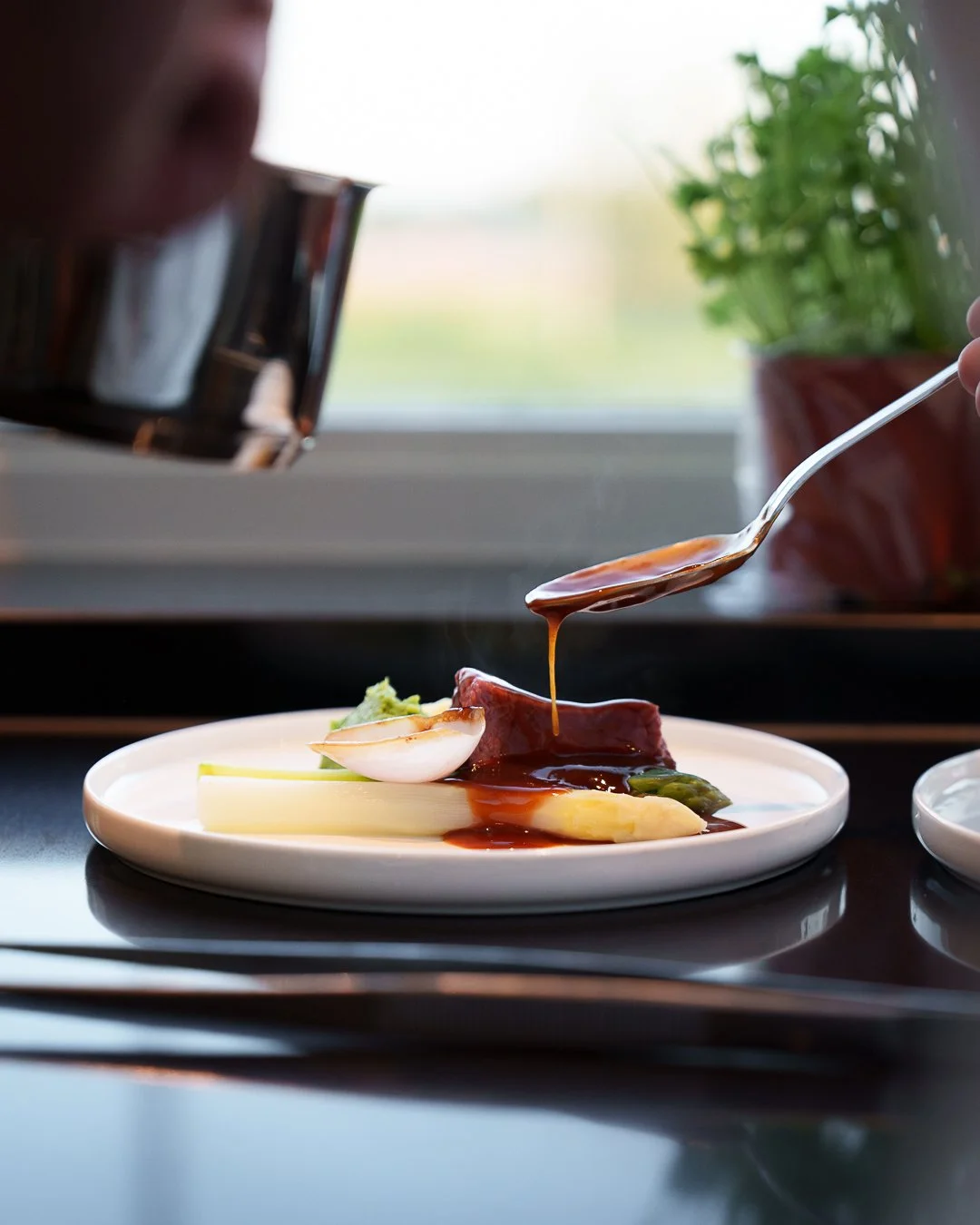 High-end gourmet dish being garnished with sauce on a white plate, featuring a piece of meat, vegetables, and a boiled egg half.