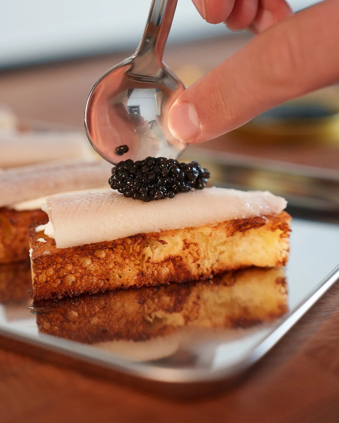 A hand placing a spoon of caviar on a slice of bread topped with a creamy spread and fish.
