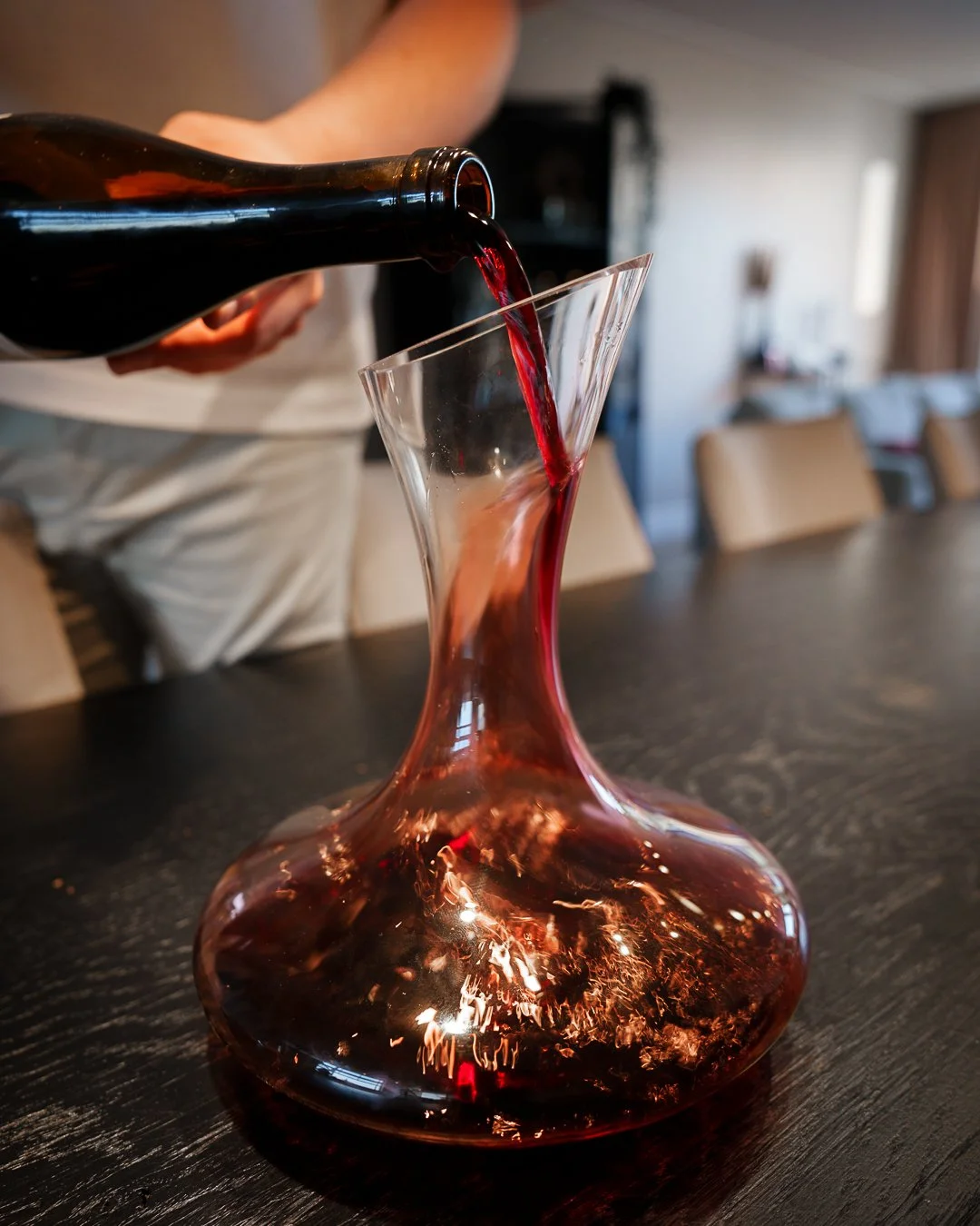 Person pouring red wine from a bottle into a glass decanter on a dark wooden table.