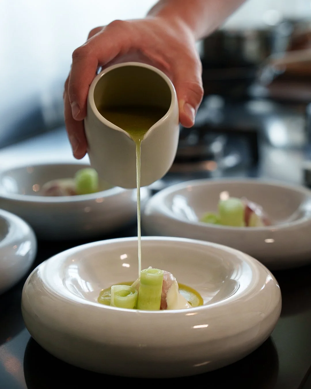 Person pouring green sauce onto gourmet dish with vegetables in white bowl.