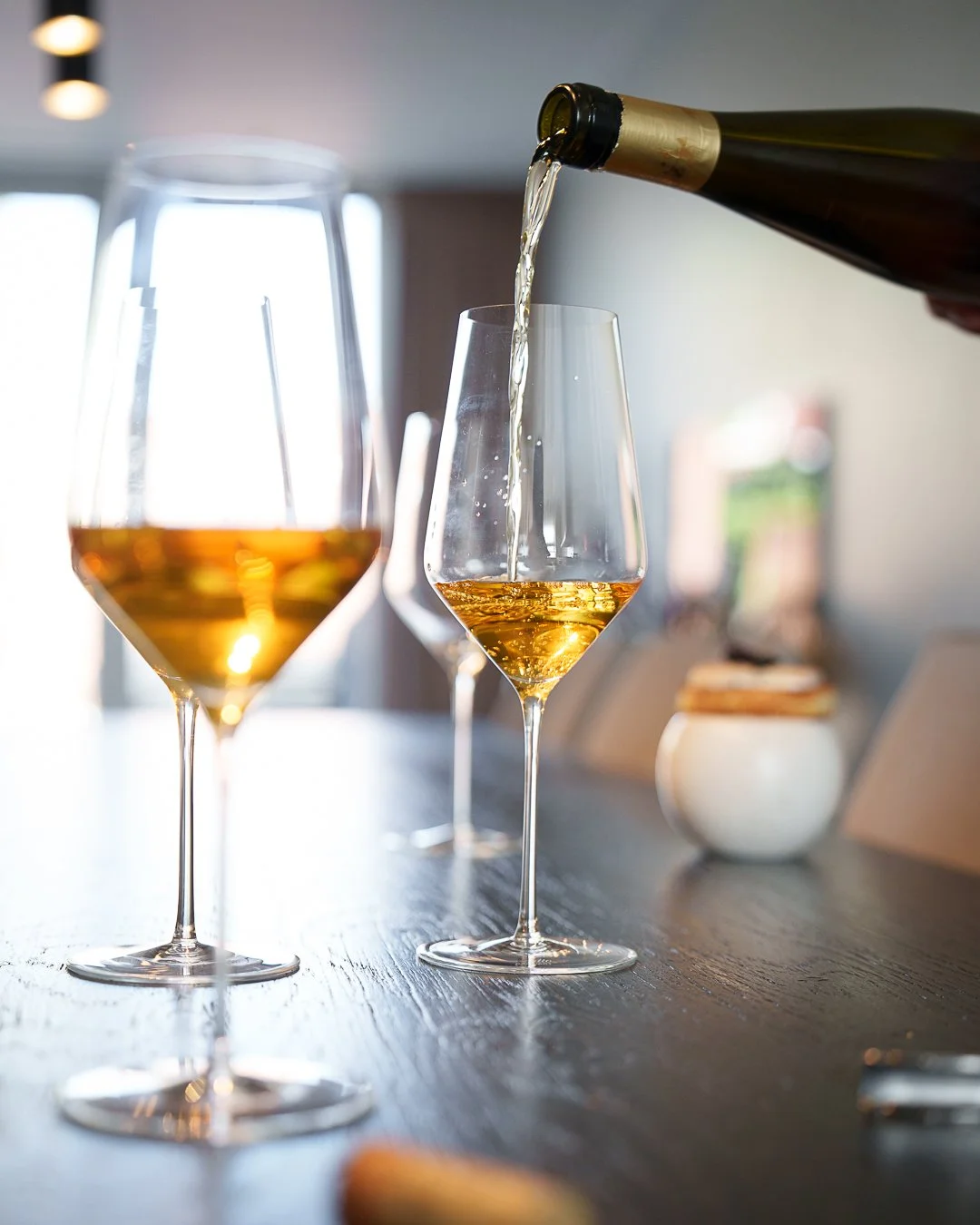 Wine being poured into a glass, with another filled wine glass in the foreground on a table.