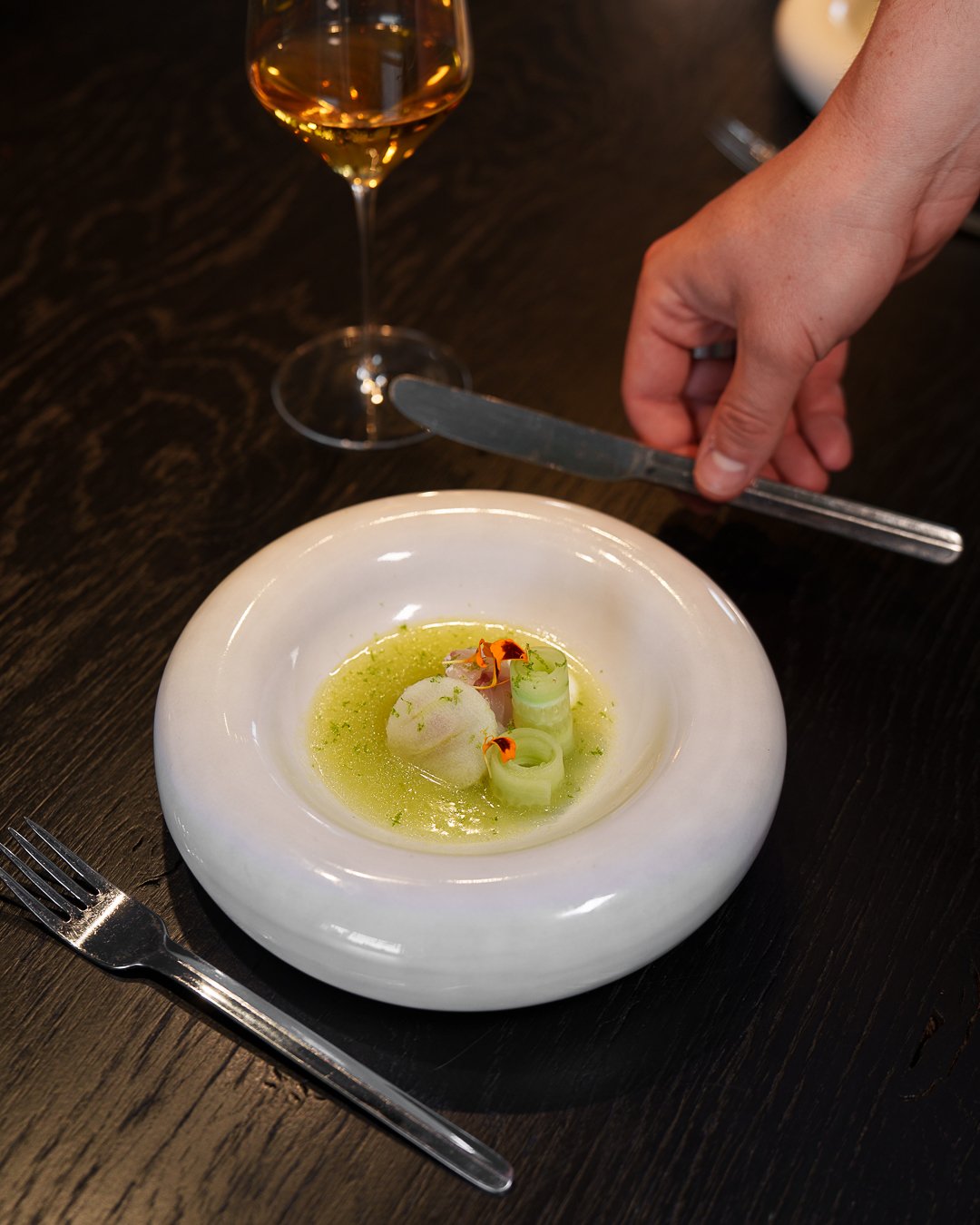 Gourmet scallop dish with cucumber, presented in a white bowl, paired with a glass of white wine. A hand is holding a knife beside the bowl, with a fork placed on the table.