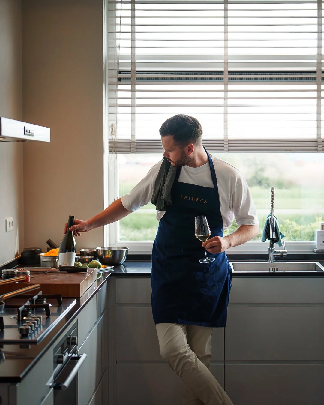 A man in a kitchen wearing an apron, holding a glass of wine and reaching for a wine bottle, with a towel over his shoulder.