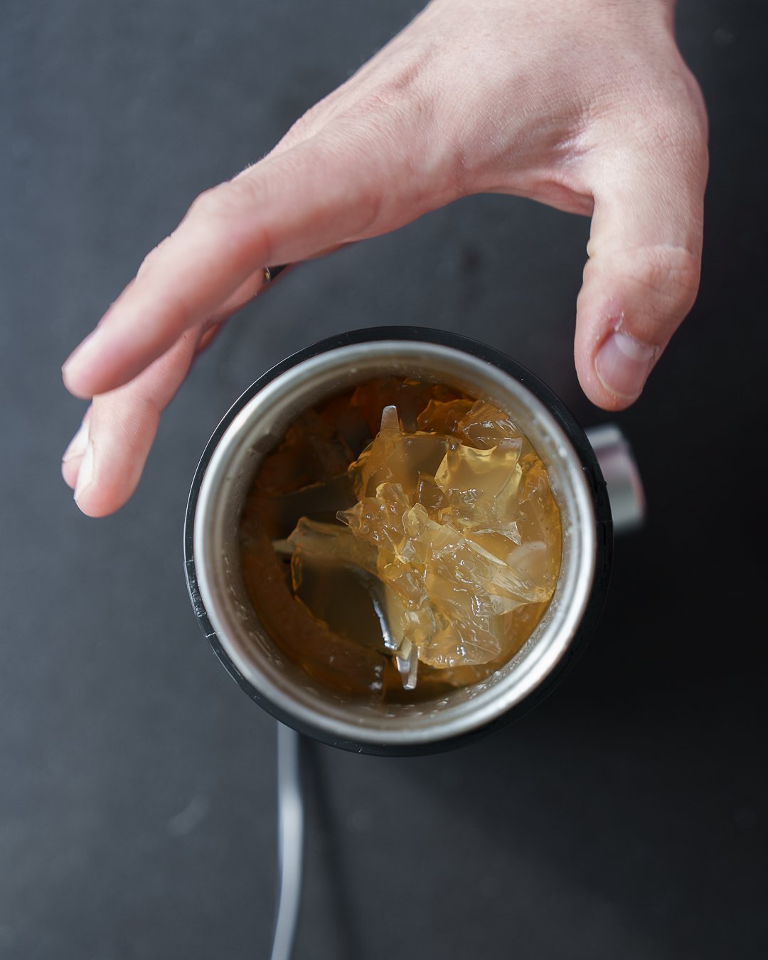 Hand holding a glass with a beverage and ice cubes on a dark surface.