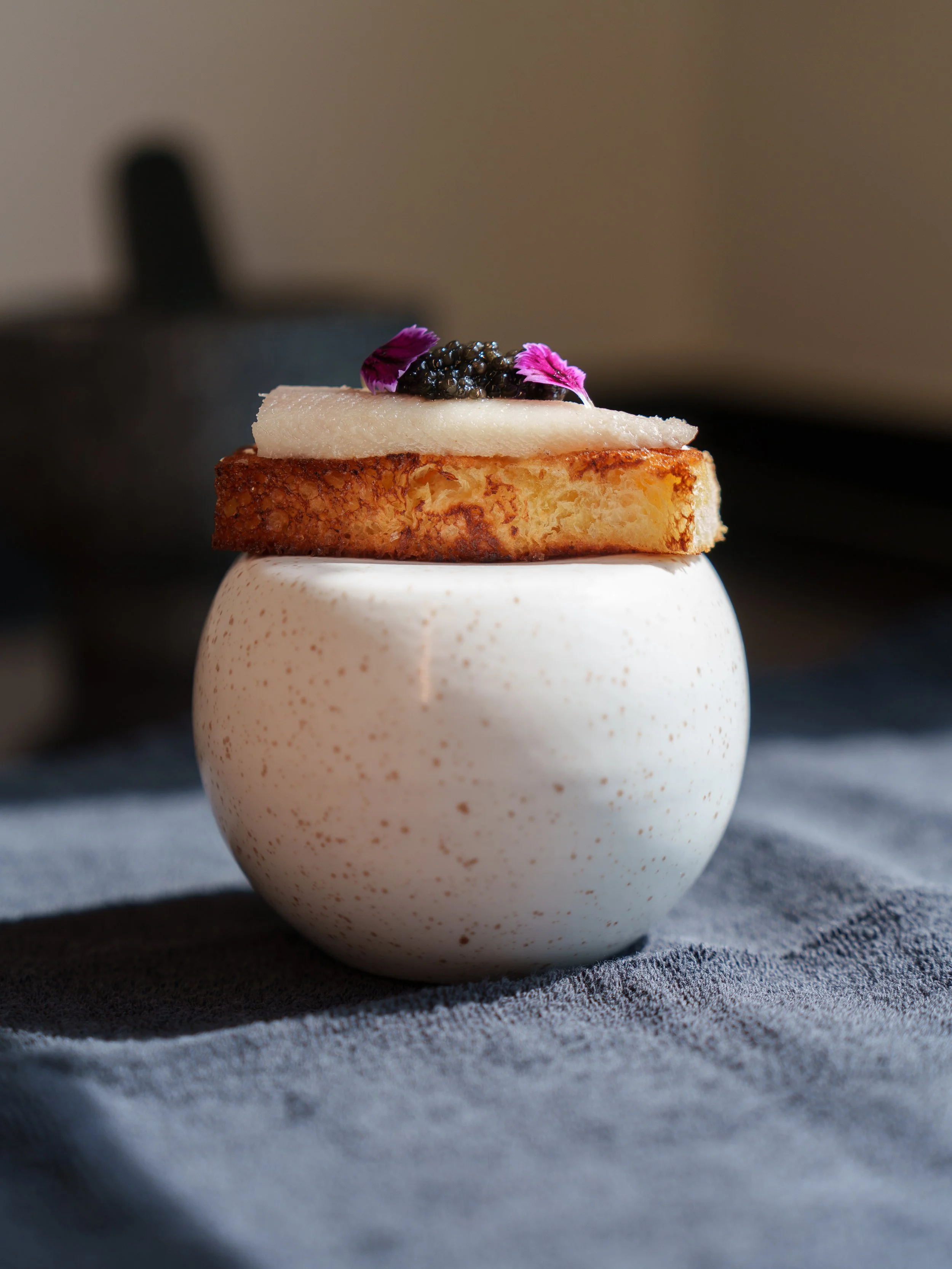 Gourmet appetizer with caviar on toast and decorative edible petals on a speckled ceramic cup.