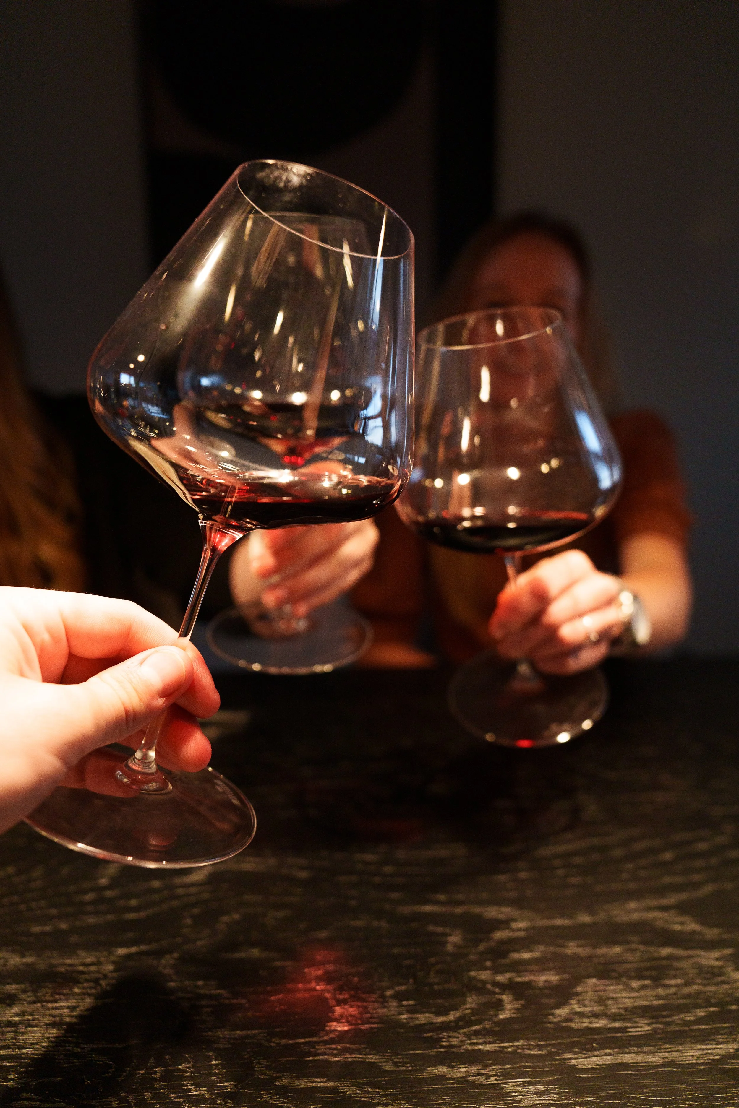 People toasting with red wine glasses, with one person in focus and others blurred in the background, sitting at a dark wooden table.