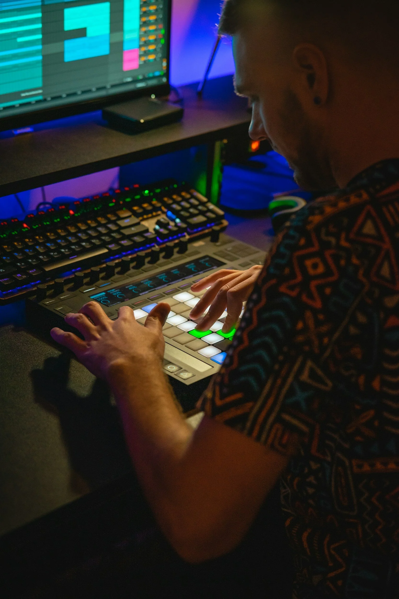 A person using a music production controller with illuminated pads, in a dimly lit room with audio equipment and a computer monitor displaying digital audio workstation tracks.