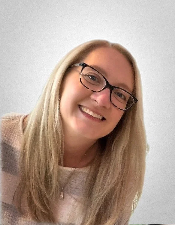A smiling woman with long blonde hair and glasses, wearing a beige top, against a plain light wall background.
