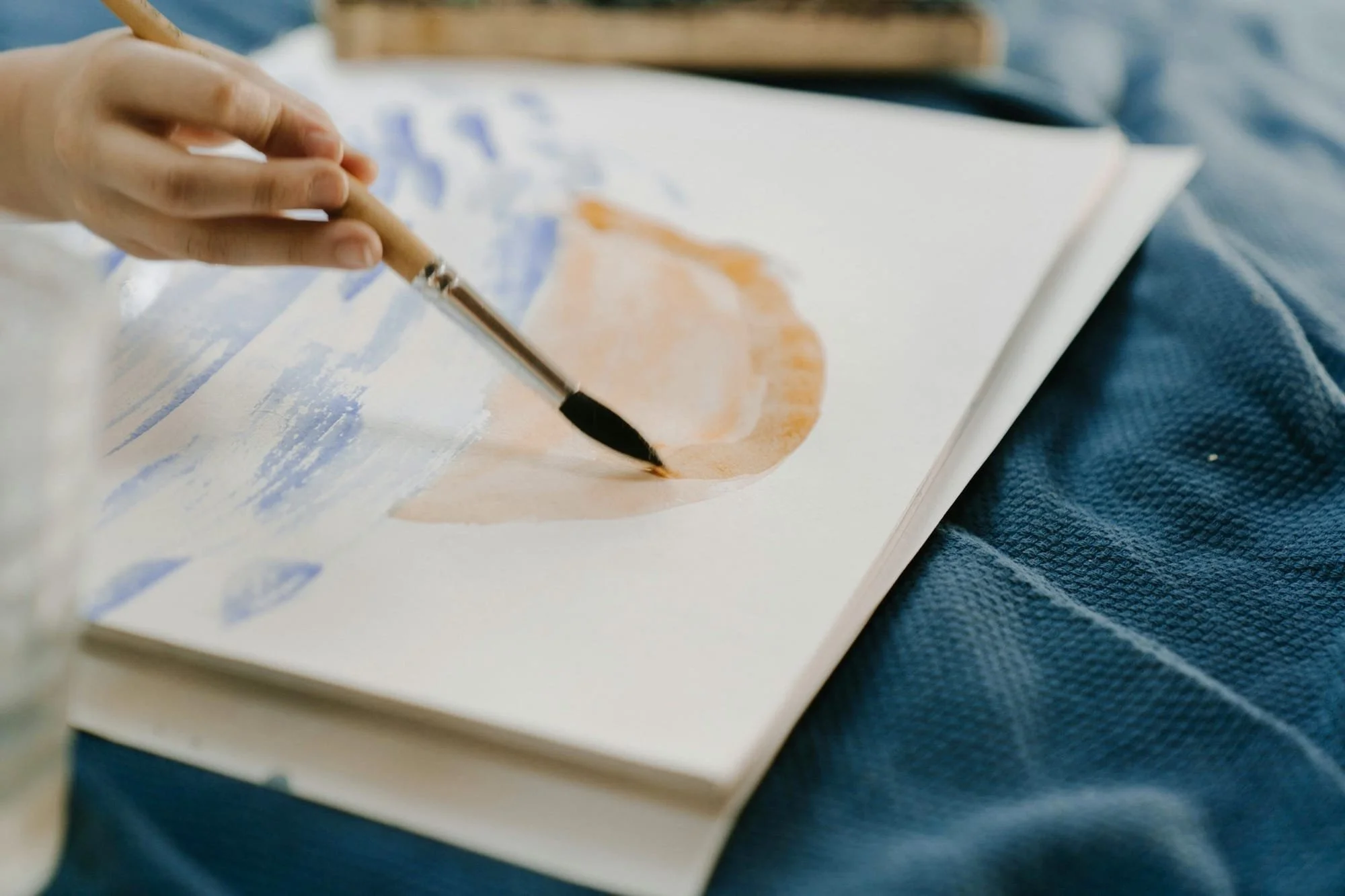 A person watercolor painting a peach on a sketchbook with a brush, resting on a blue textured fabric.