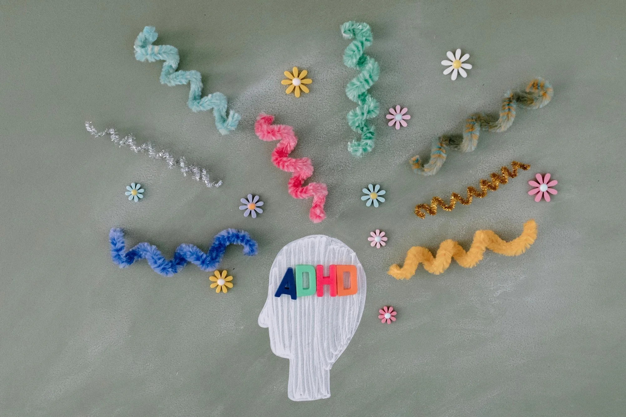 Paper cut-out of a human head in profile with colorful magnetic letters spelling 'ADHD' on the forehead, surrounded by various colored pipe cleaners and small flower-shaped decorations on a gray background.