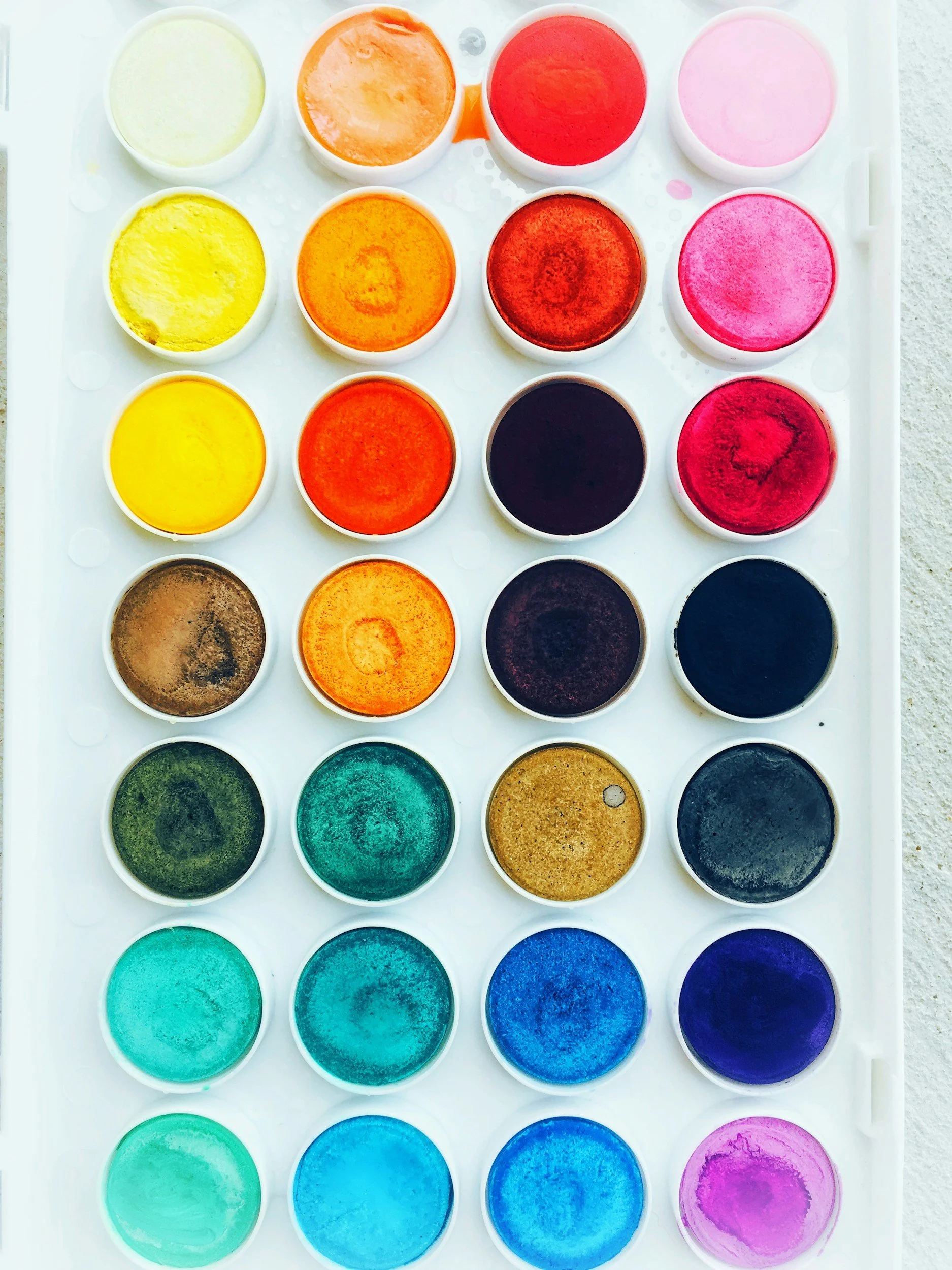 A watercolor paint set with 24 circular pans of various colors, including yellow, orange, red, pink, black, brown, green, blue, and purple, arranged on a white tray.