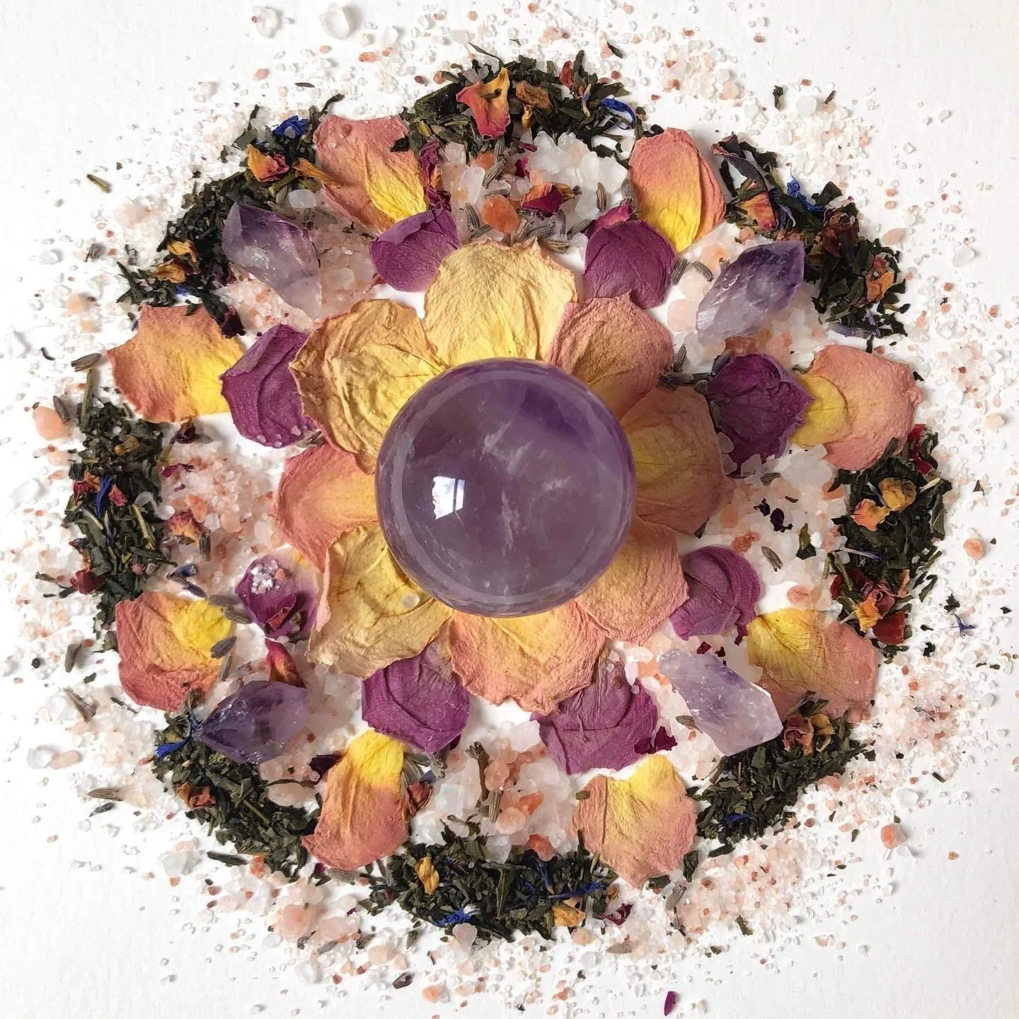 A circular arrangement of dried rose petals, herbs, and salt around a purple crystal ball on a white surface.