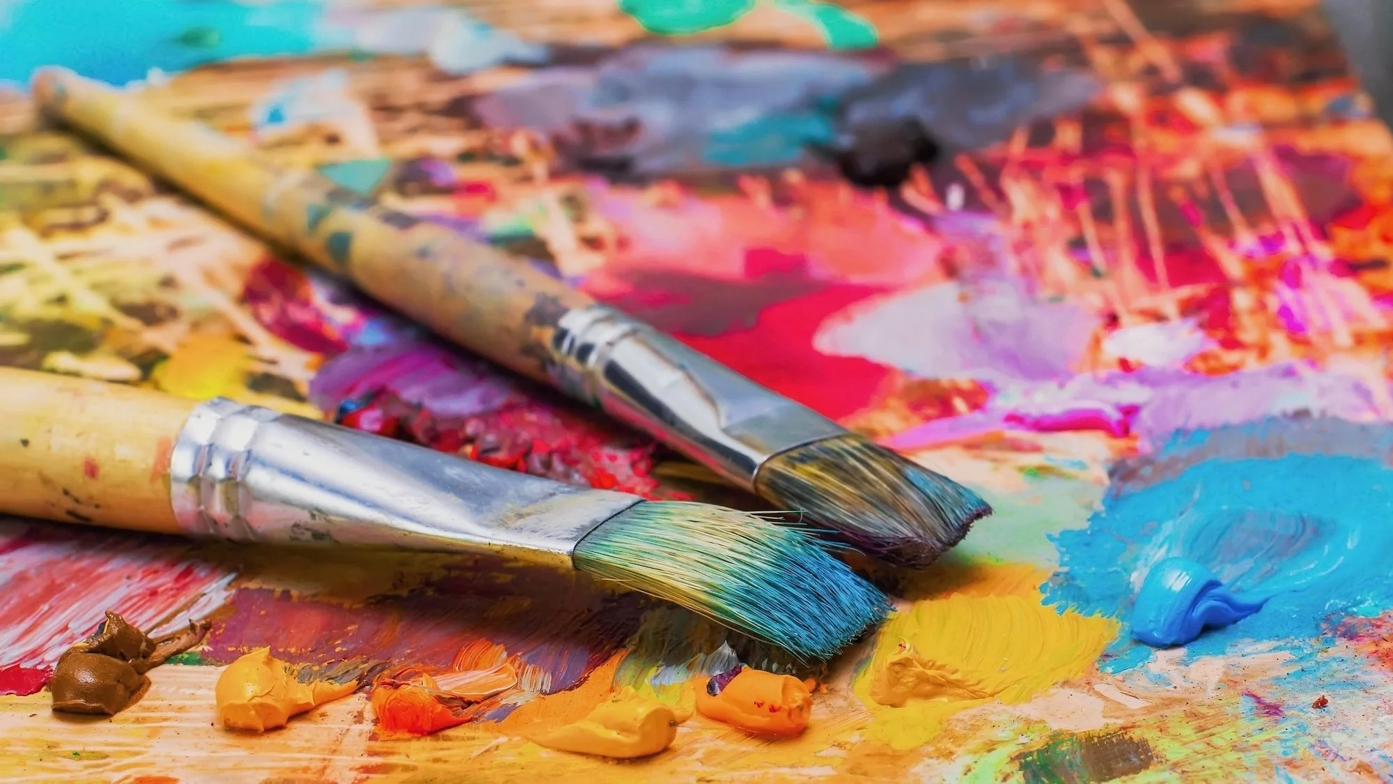A colorful artist's palette with various paint blobs and two paintbrushes resting on it, surrounded by mixed and smeared bright and pastel paint colors.