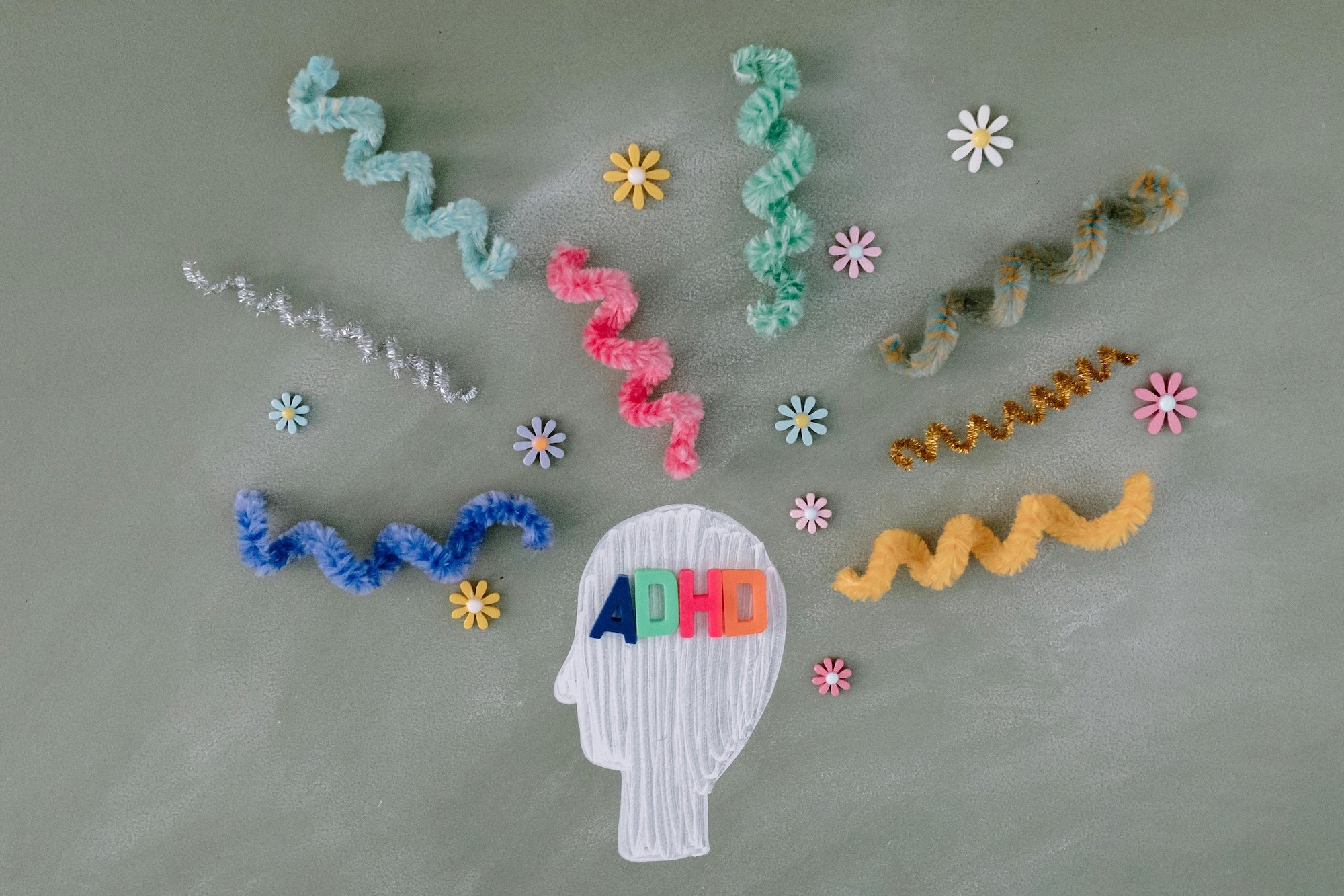Decorative chalk drawing of a human head silhouette with colorful pipe cleaners and small flower cutouts around it, with the word 'ADHD' in multicolored letters across the head.