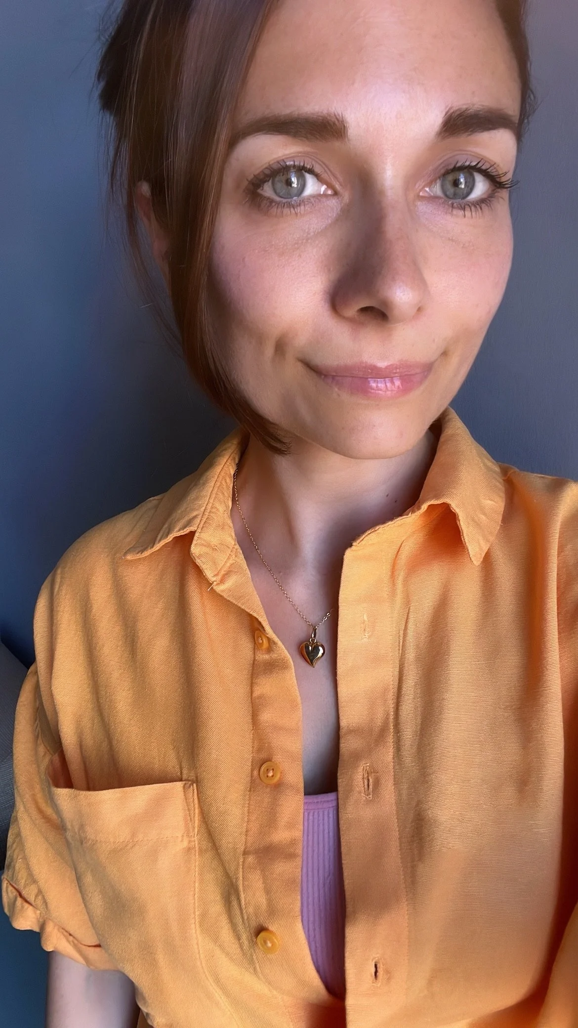 A young woman with short brown hair and blue eyes wearing a yellow button-up shirt and a necklace with a heart-shaped pendant.