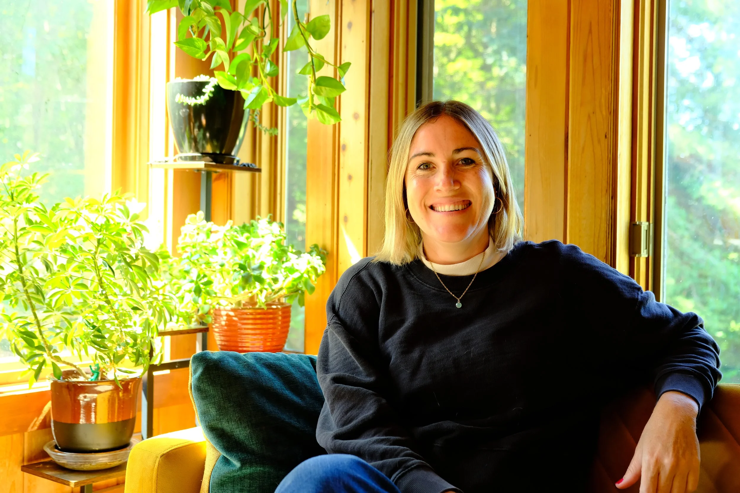 A woman with shoulder-length blonde hair smiling and sitting in a room with large windows, surrounded by green plants and natural wood decor.