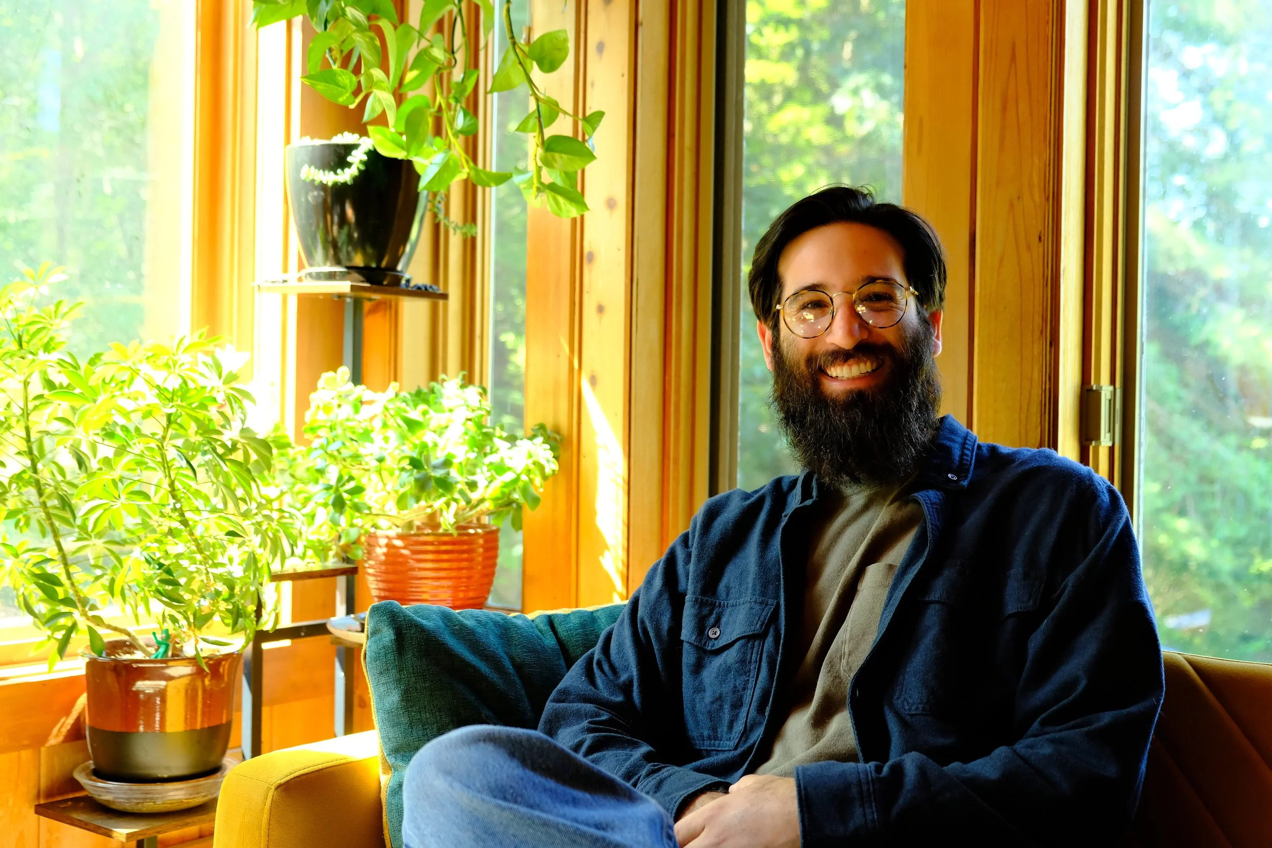 A smiling man with a beard and glasses sitting indoors near a large window of a wooden room, surrounded by green plants on a shelf and a table.