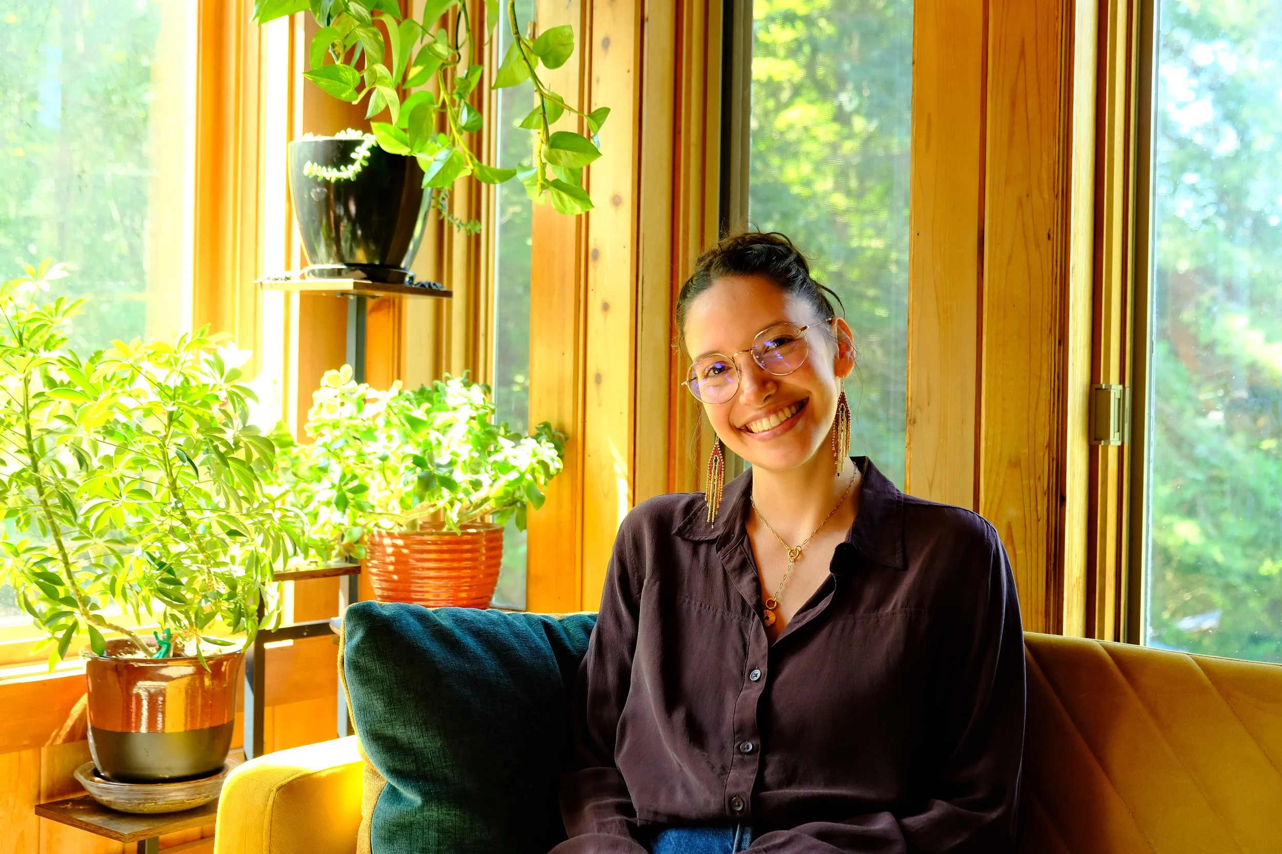 A woman with glasses smiling, seated in a sunlit room with wooden walls, surrounded by green plants, with a collection of windows showing trees outside.