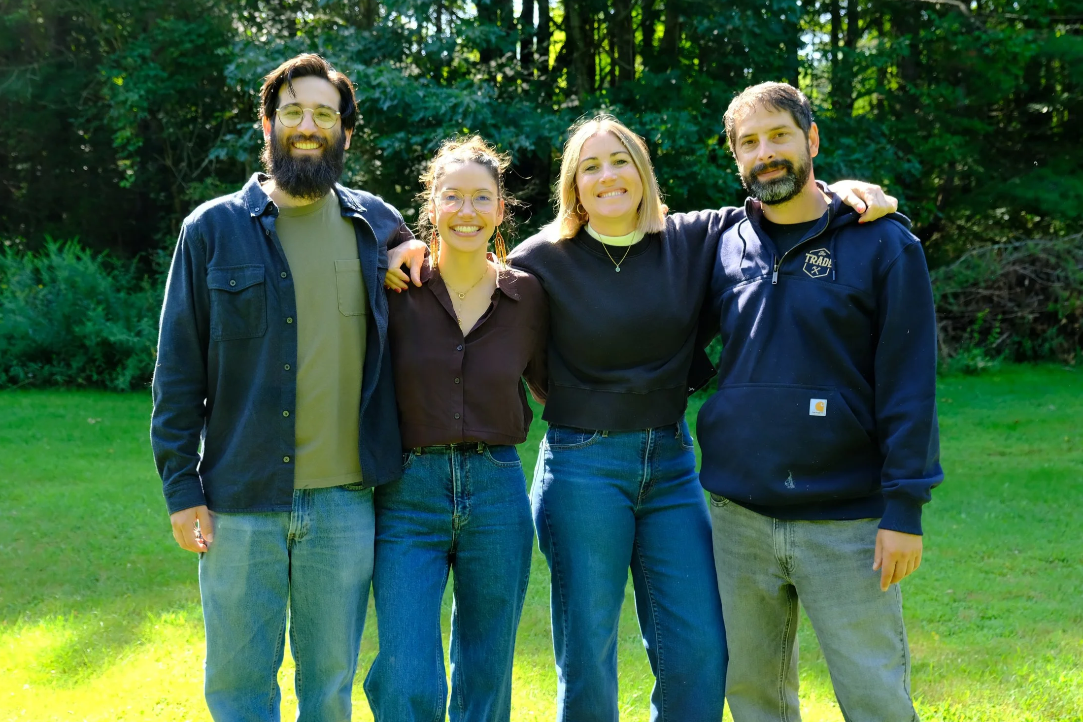 A group of four friends standing outdoors on grass, smiling and posing for the camera with a background of green trees.