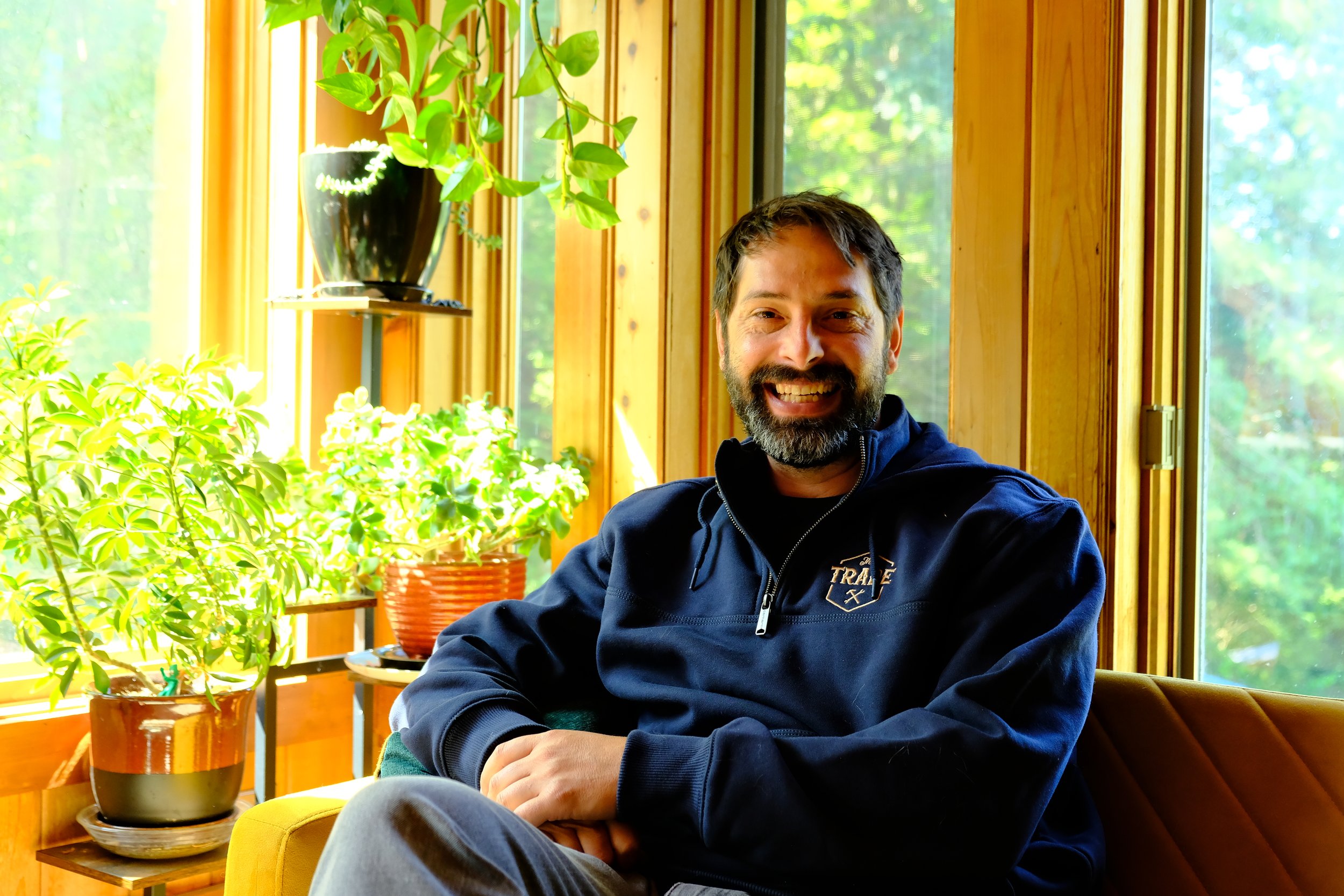 A smiling man with dark hair and a beard sitting indoors near a window with wooden framing, surrounded by green potted plants.