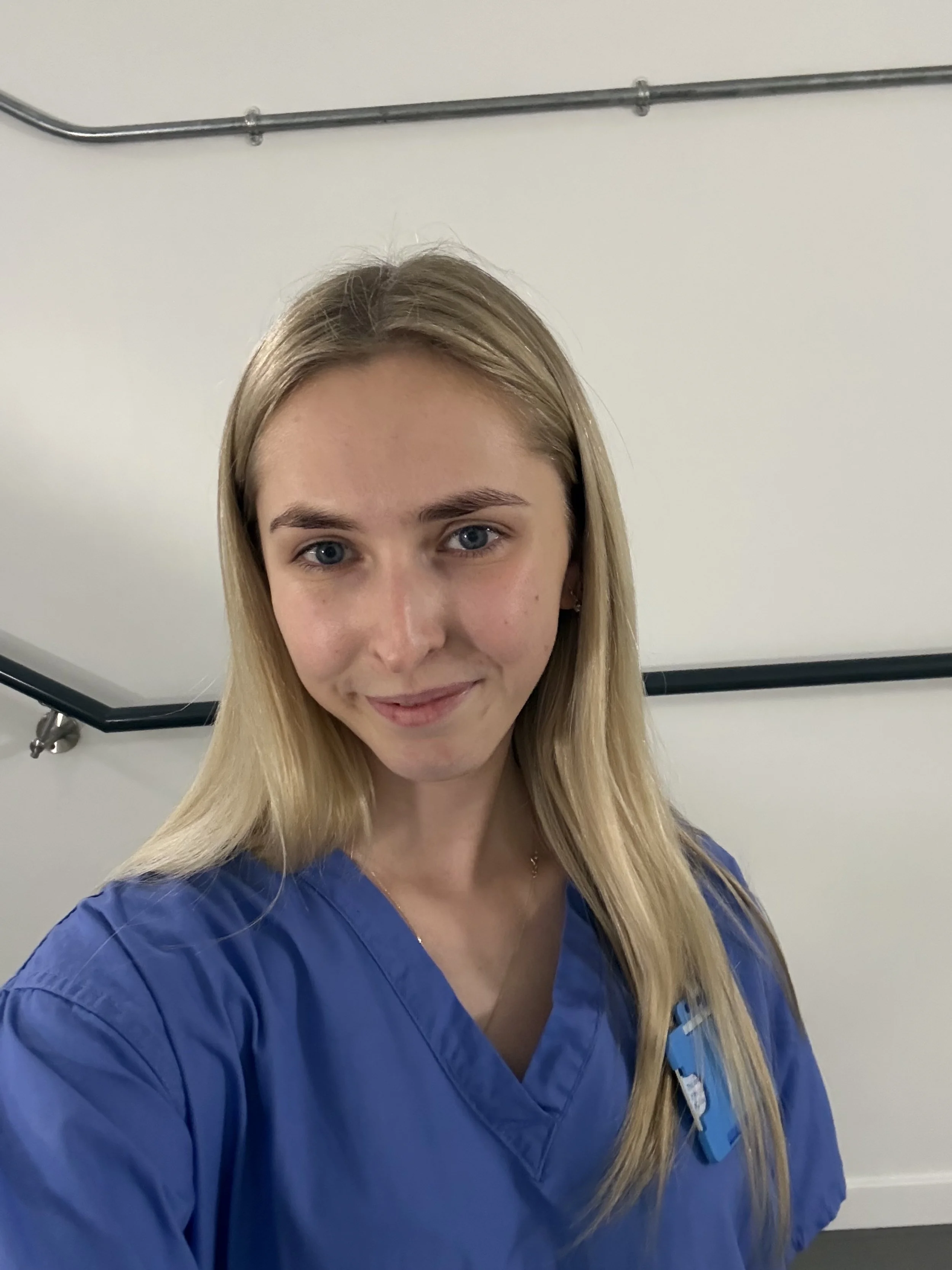A young woman with long blonde hair and blue eyes wearing blue medical scrubs, standing near a white wall with a handrail and metal pipe overhead.