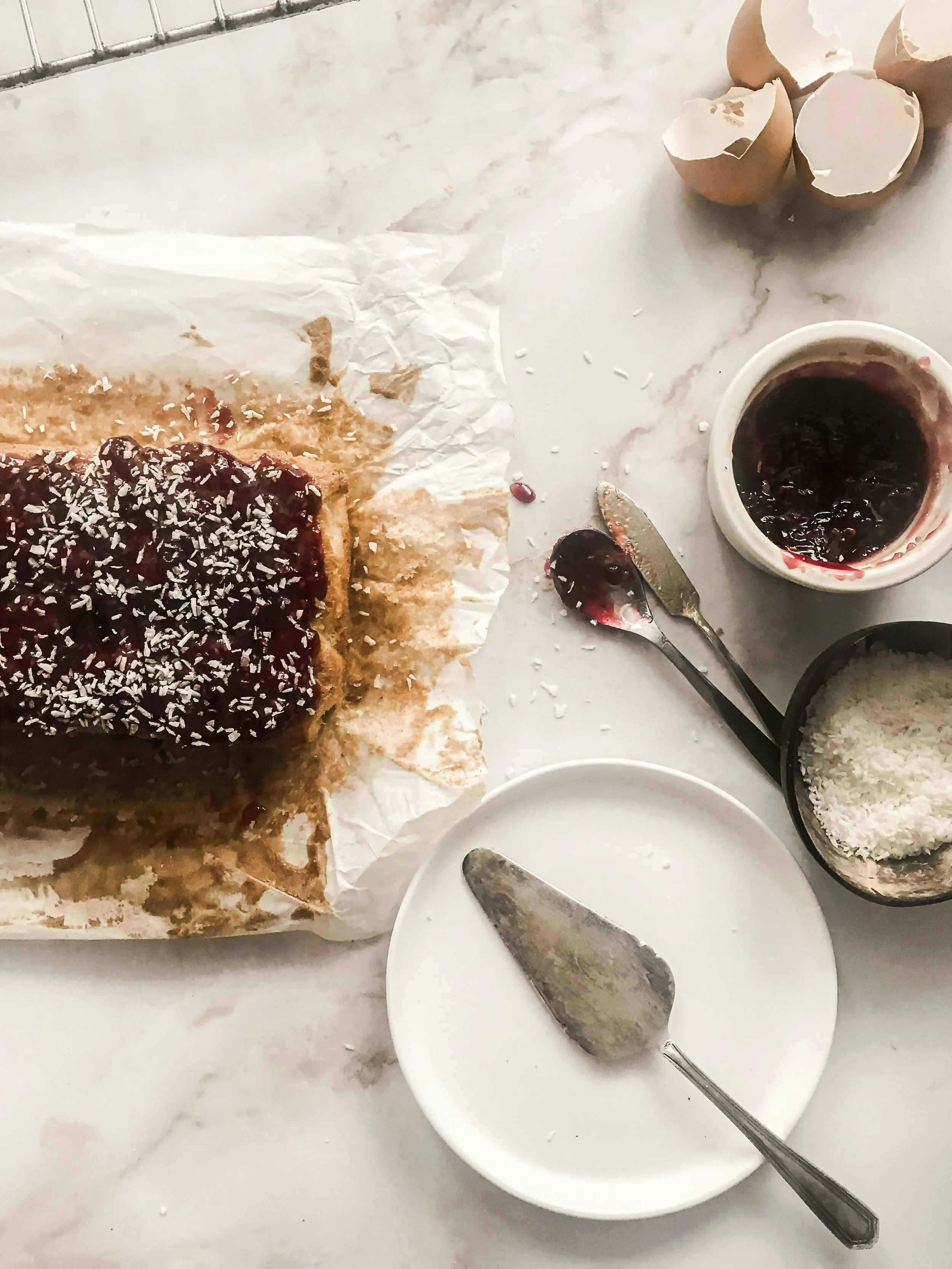 A partially eaten coconut cake with a cherry topping and shredded coconut on parchment paper, with eggshells, a jar of cherry filling, a small bowl of shredded coconut, a metal cake server, and a small white plate on a marble surface.