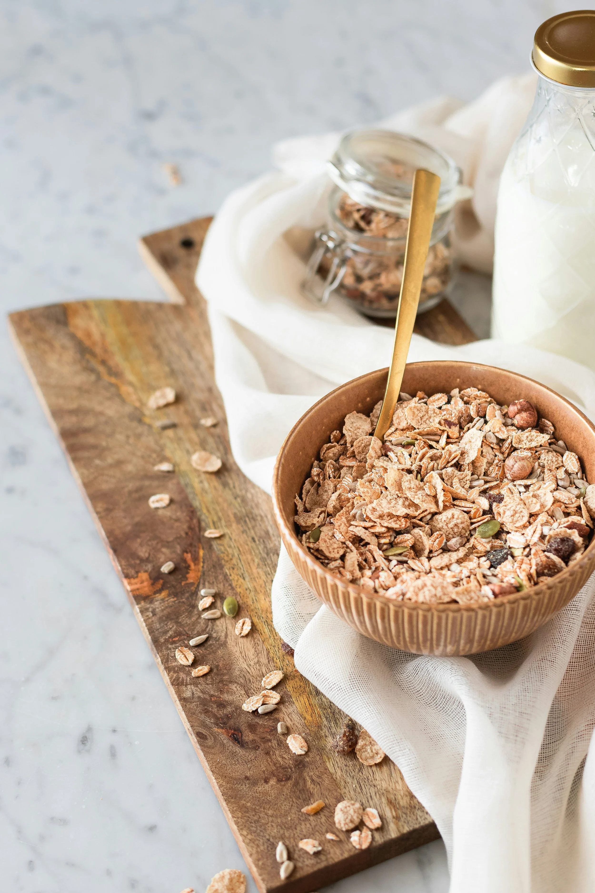 A bowl of granola with a spoon, placed on a white cloth on a wooden board, with scattered granola pieces around.