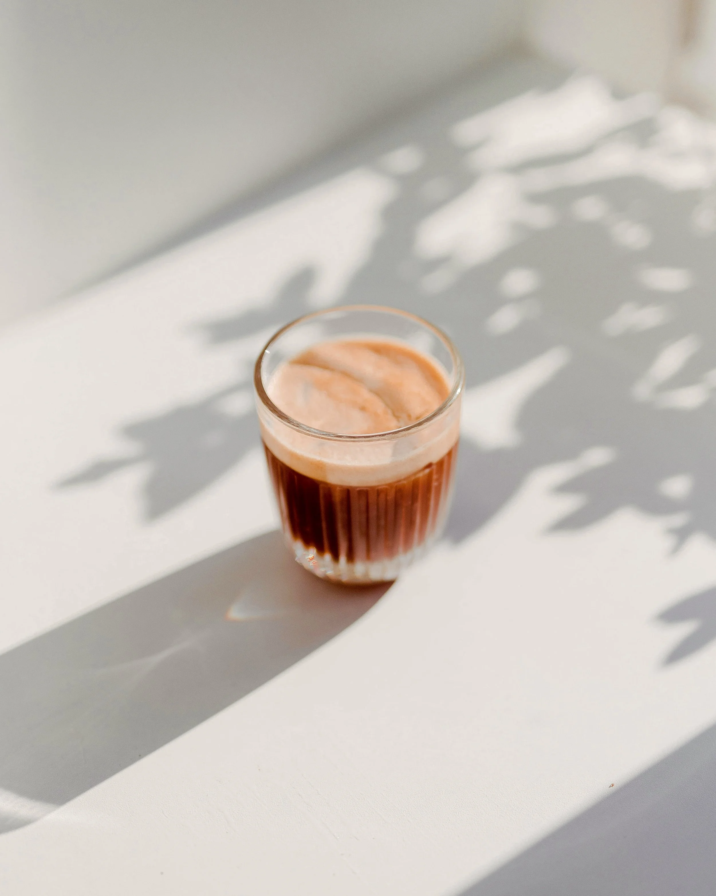 A glass of pinkish juice or smoothie on a white surface with shadows of leaves.