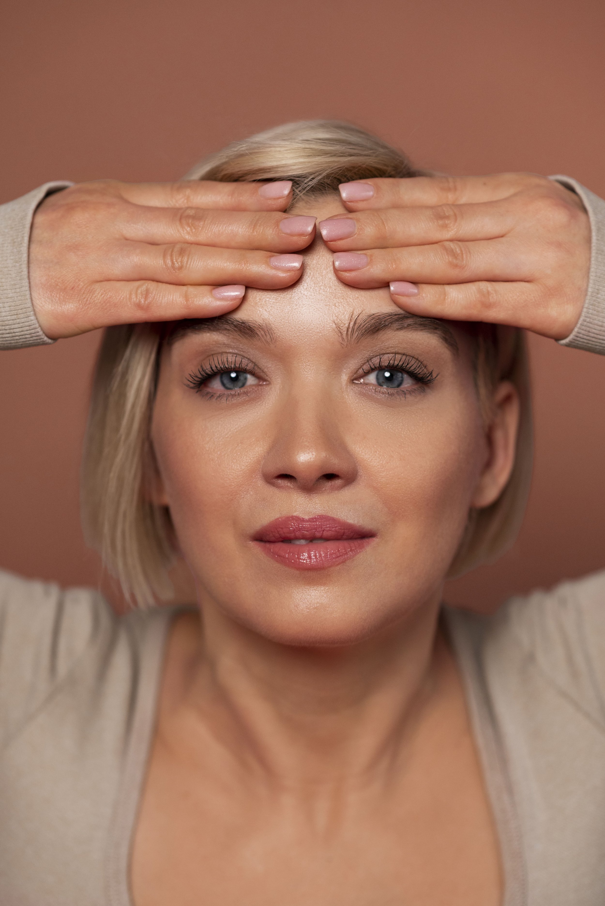 Best Adelaide Injector - SHEER MEDICAL AESTHETICS A woman with short blonde hair and blue eyes holding her forehead with both hands against a pink background. Best Adelaide Injector - SHEER MEDICAL AESTHETICS