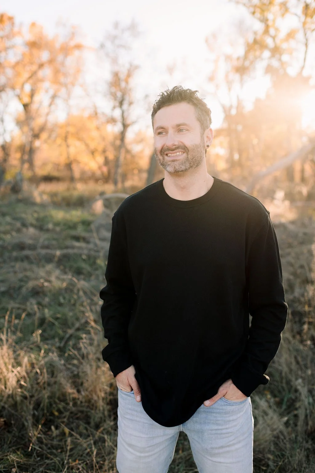 A smiling man with a beard and short curly hair stands outdoors in a park during sunset, wearing a black long-sleeve shirt and light-colored jeans.