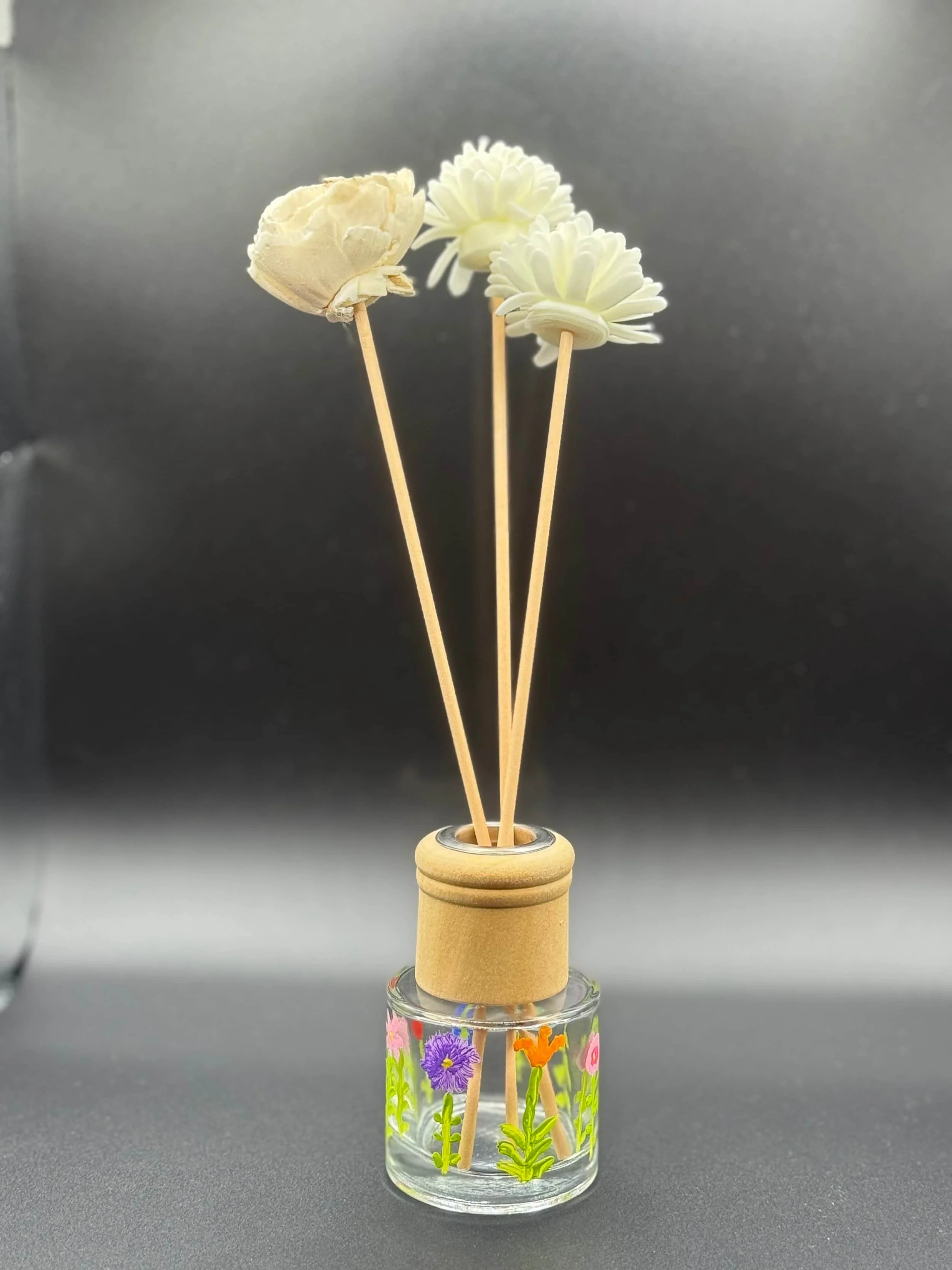A glass diffuser jar with painted flowers, holding three white artificial flowers on wooden sticks, against a black background.